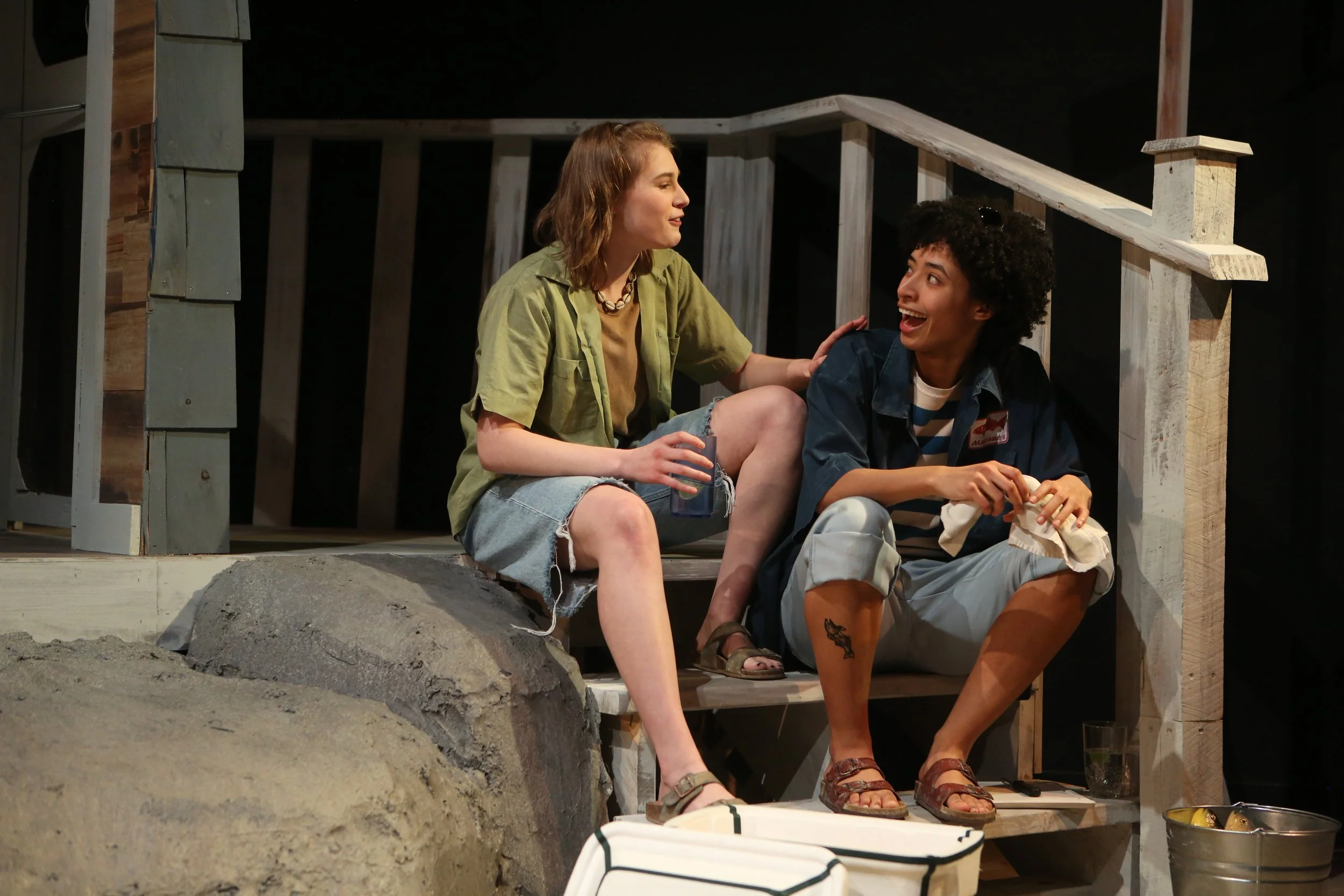 Two women sitting on the porch of a small house at night, engaged in conversation. One woman has curly hair, sunglasses, and a denim jacket, while the other has shoulder-length hair, a necklace, and a green shirt. They appear happy and relaxed.