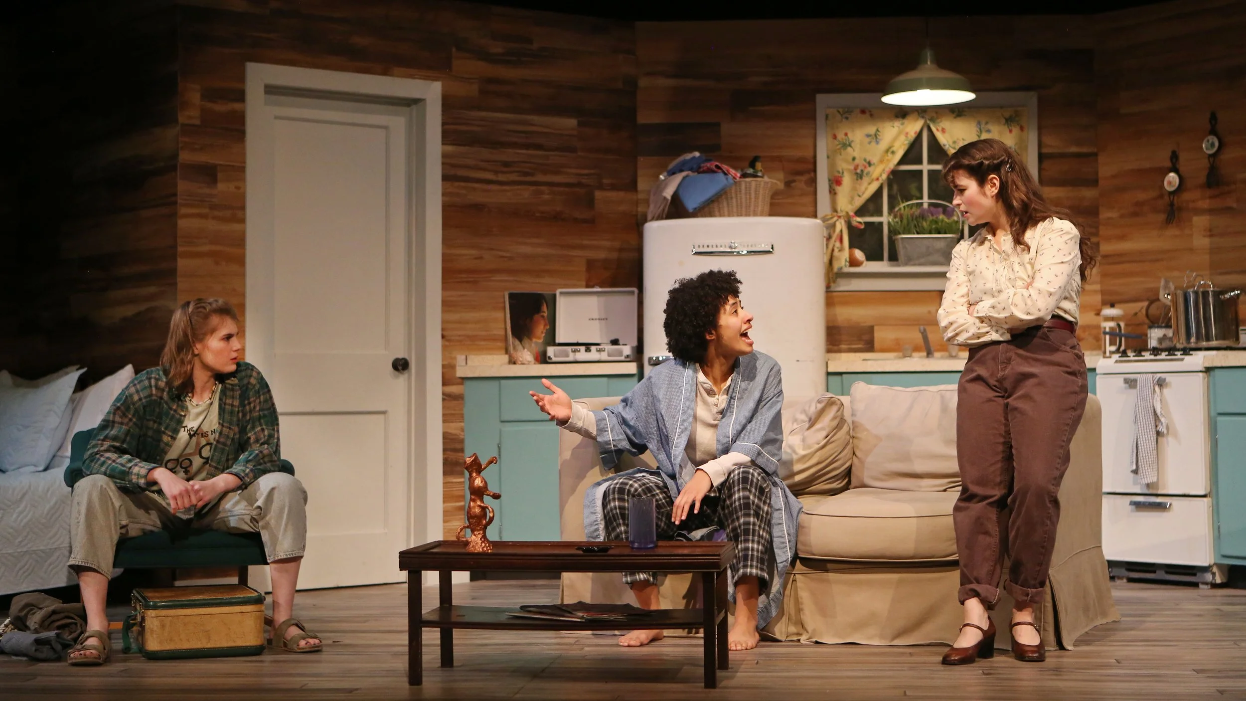 Four women are in a living room with wood-paneled walls, engaged in conversation. One woman is sitting on a couch, another on a chair with a suitcase, a third is kneeling on the floor, and the fourth is standing, emotional. The room has a vintage fri