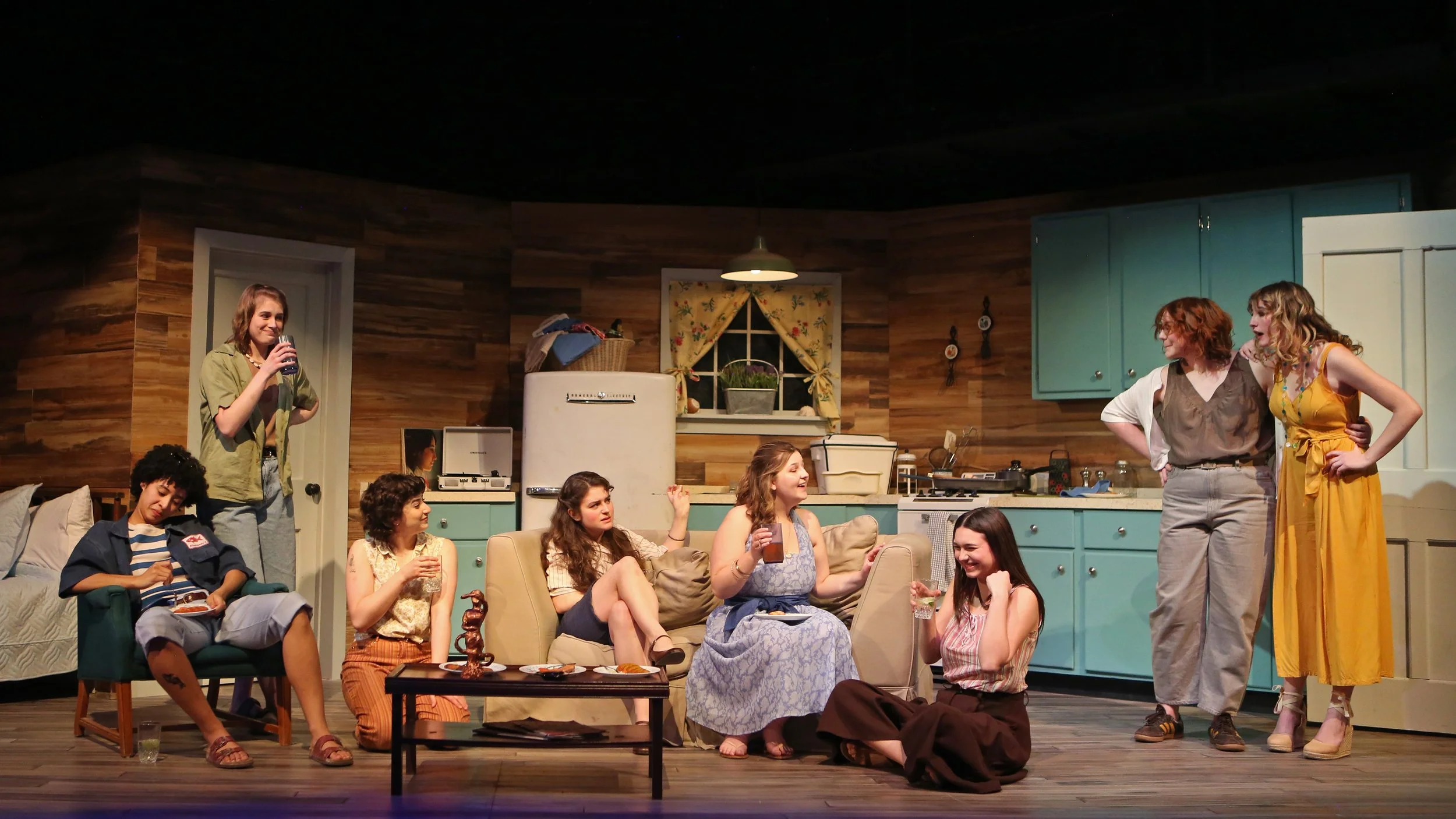 A play with multiple actors on stage in a cozy, retro-style kitchen set, engaging in dialogue and socializing.