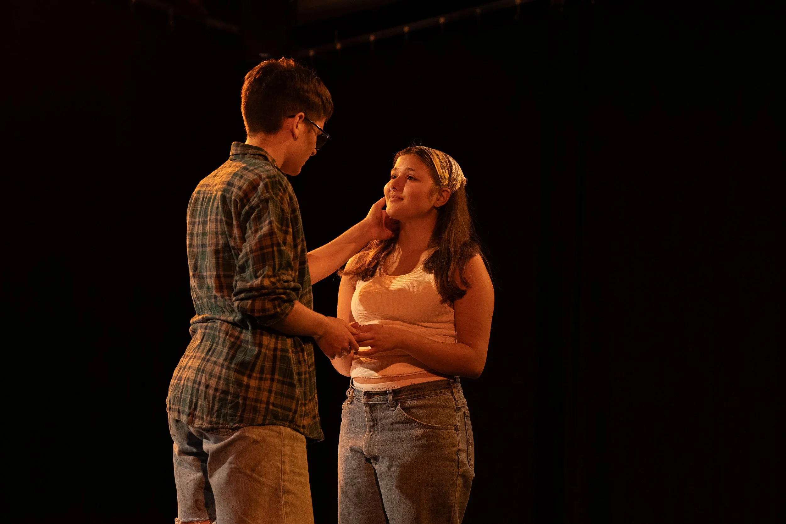 A young man in a plaid shirt and a young woman in a tank top and high-waisted jeans are on stage, holding hands and looking at each other, with the young man's other hand gently touching the woman's face.
