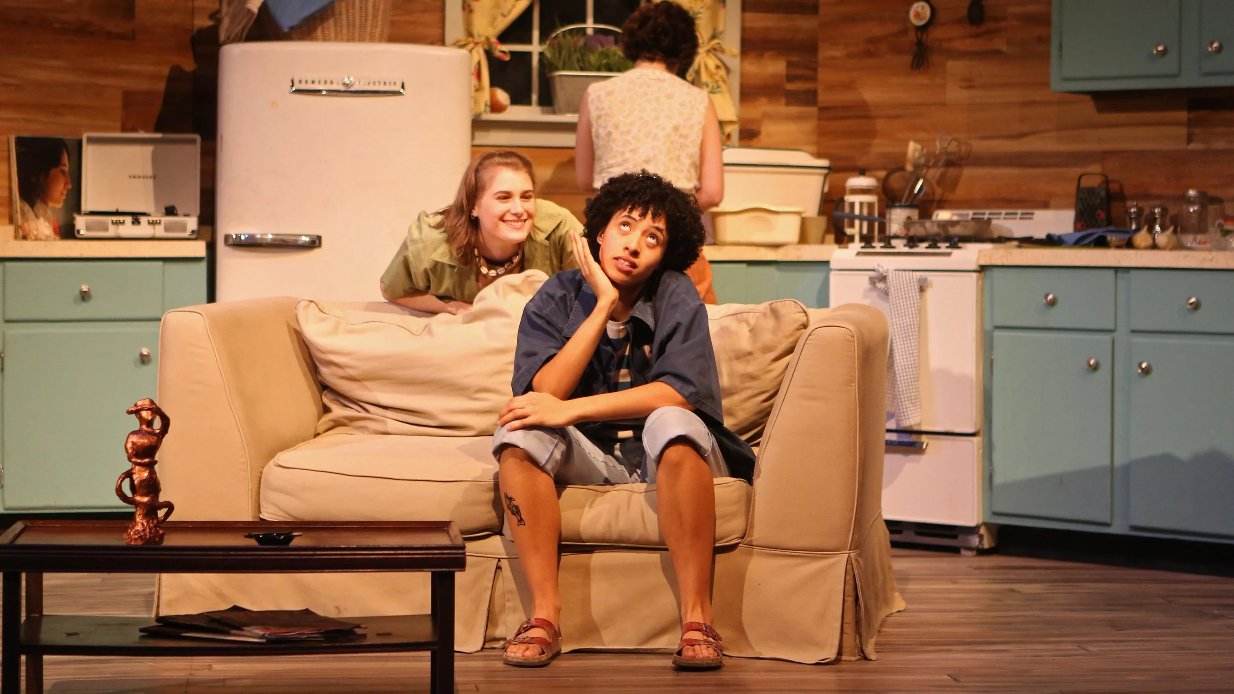 Two women and one man in a kitchen, with the women smiling and the man sitting on a sofa with a thoughtful expression. The kitchen has teal cabinets, a white retro refrigerator, and wooden walls.