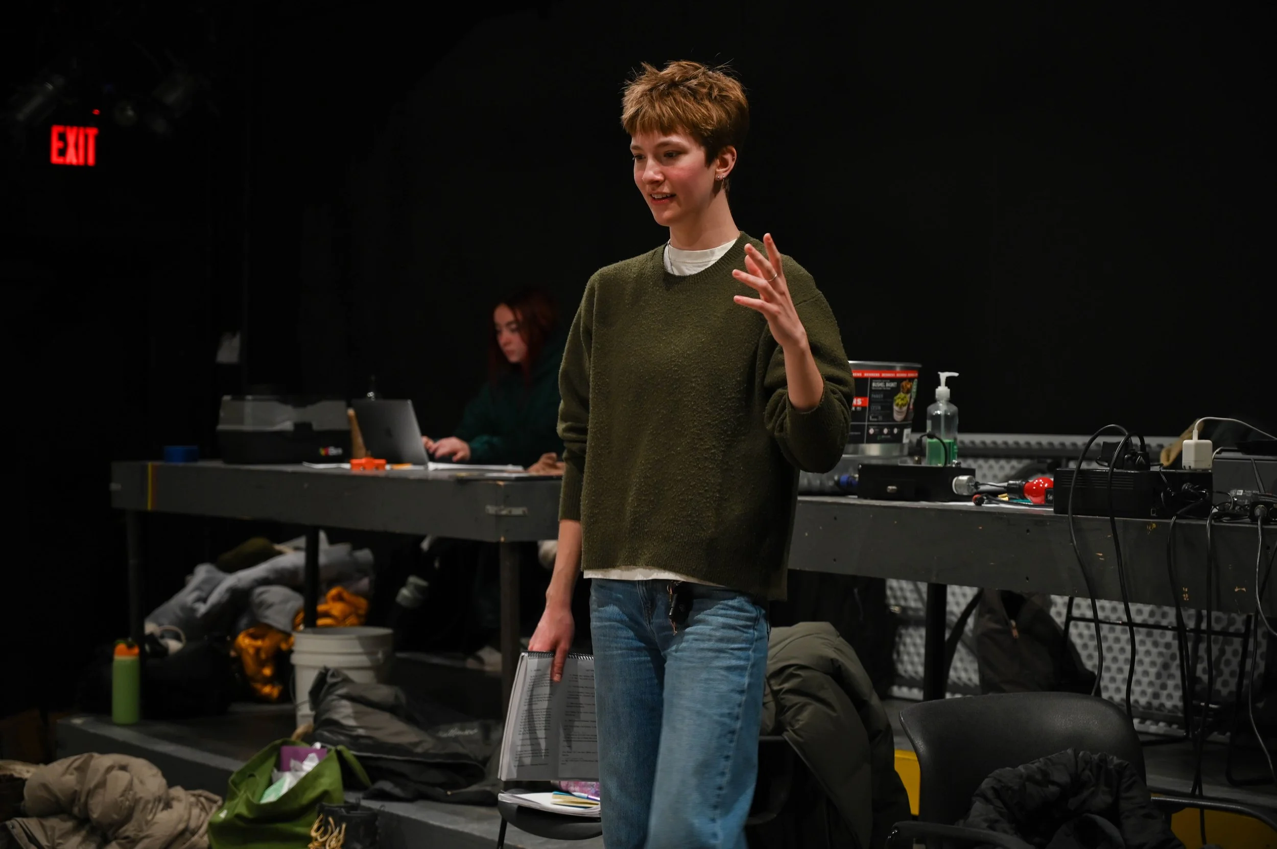 Young woman with short hair, wearing a green sweater and jeans, speaking or gesturing in a dark room with people and equipment in the background.