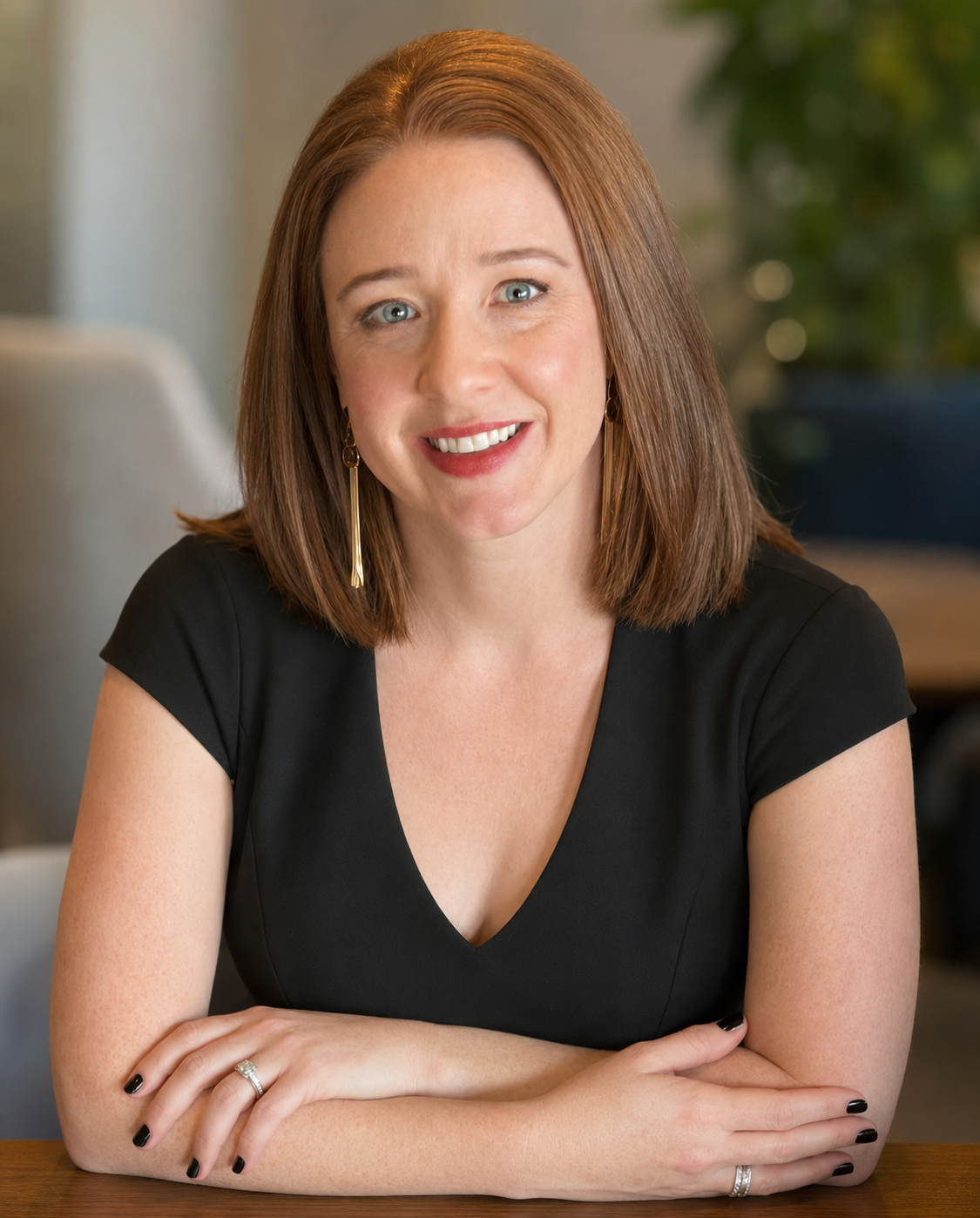 A woman with shoulder-length reddish-brown hair, wearing a black V-neck top and gold earrings, smiling and resting her arms on a table.