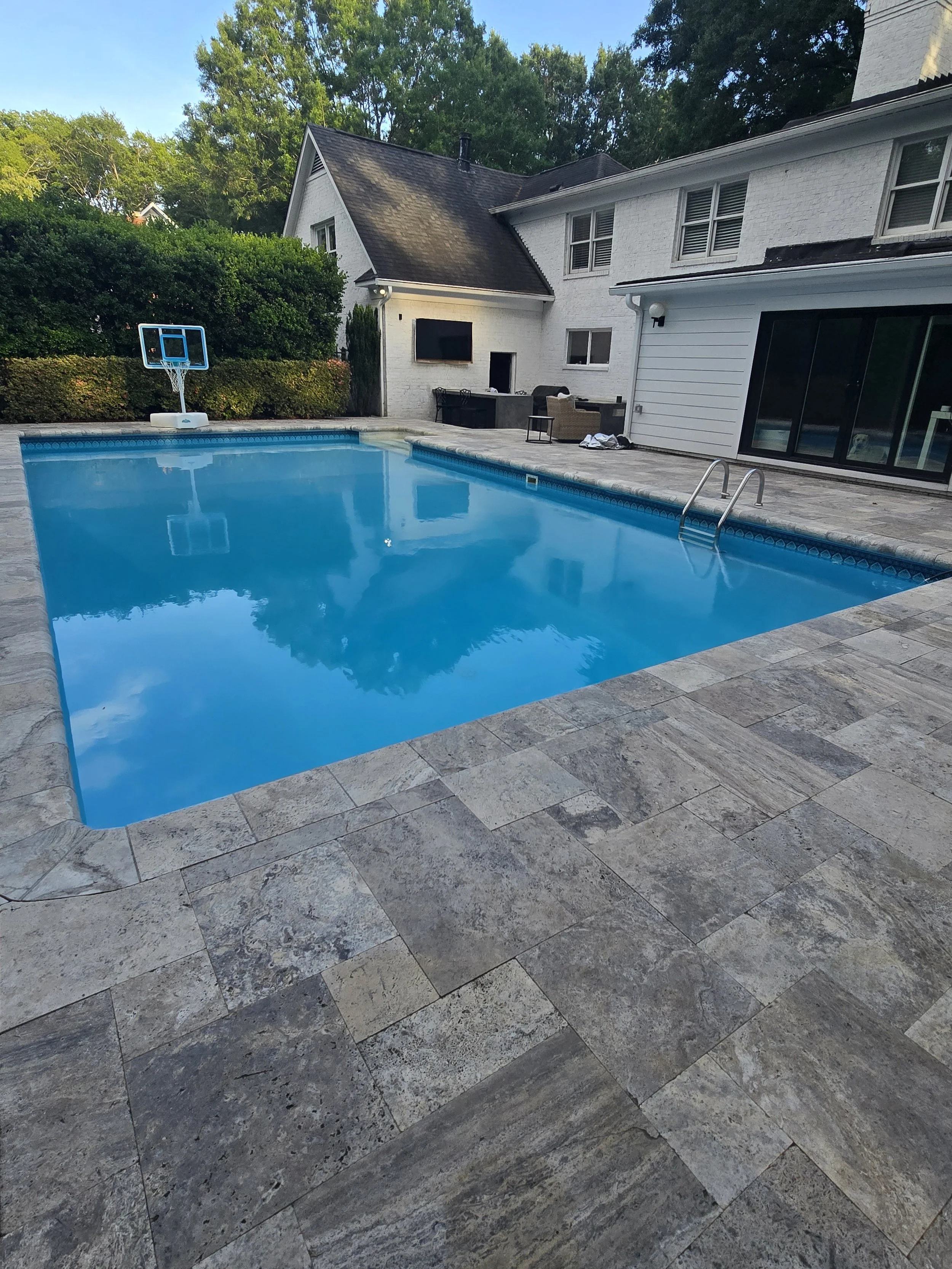 Backyard with a swimming pool serviced by AB Premium Aqua Solutions, outdoor furniture, a basketball hoop, and a house with white brick and siding, surrounded by trees.