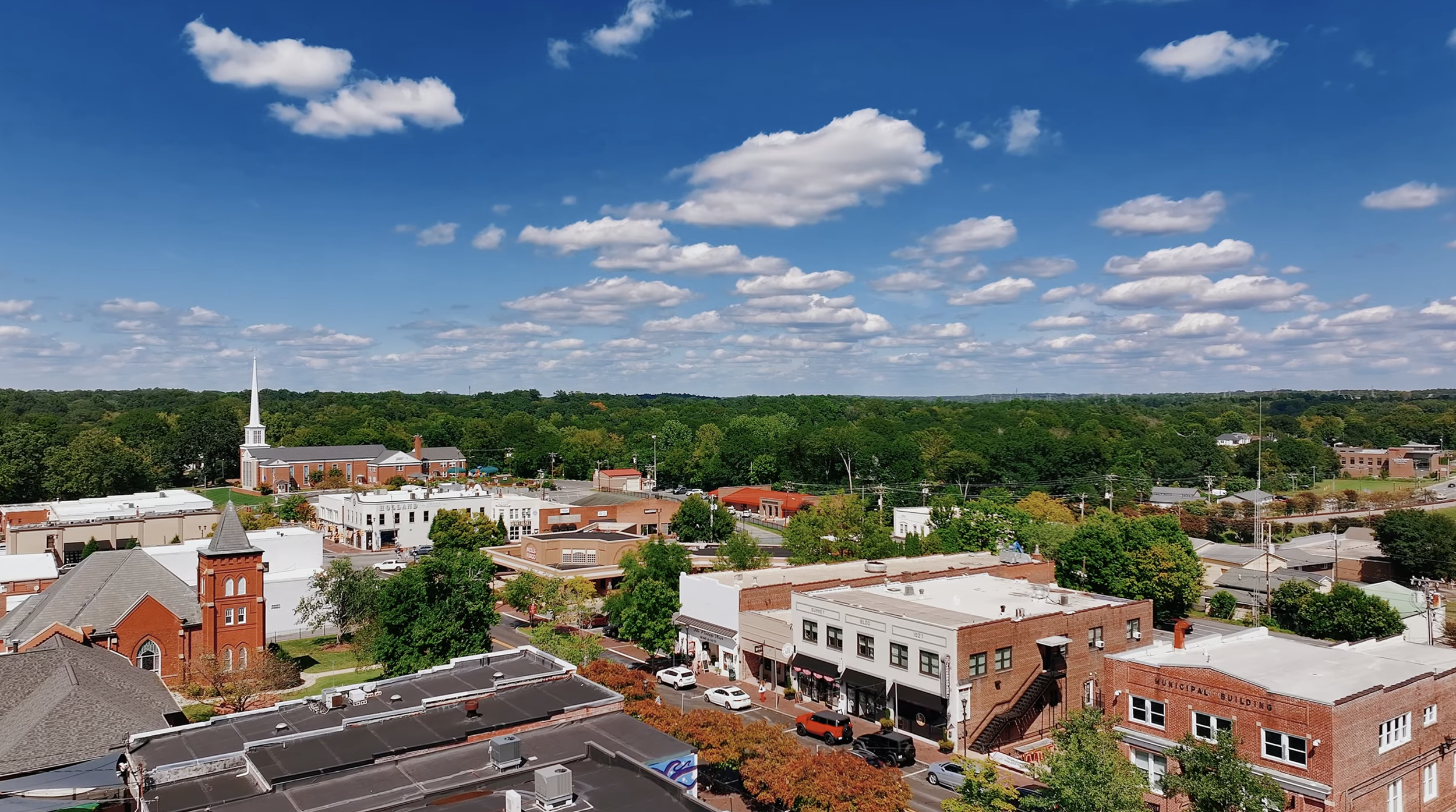 Aerial view of a small town with a church, brick buildings, trees, and a bright blue sky with fluffy white clouds.