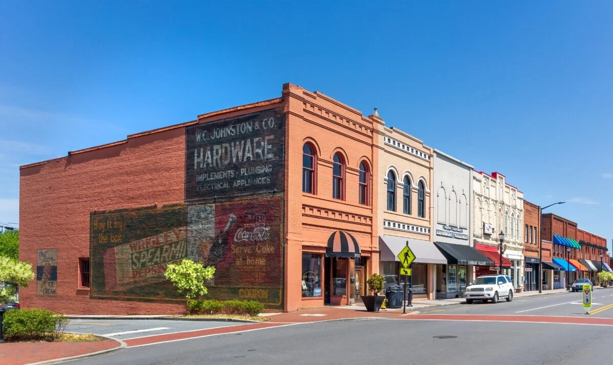A row of historic brick buildings on a small-town street under a bright blue sky, with storefronts, awnings, and cars parked along the curb.