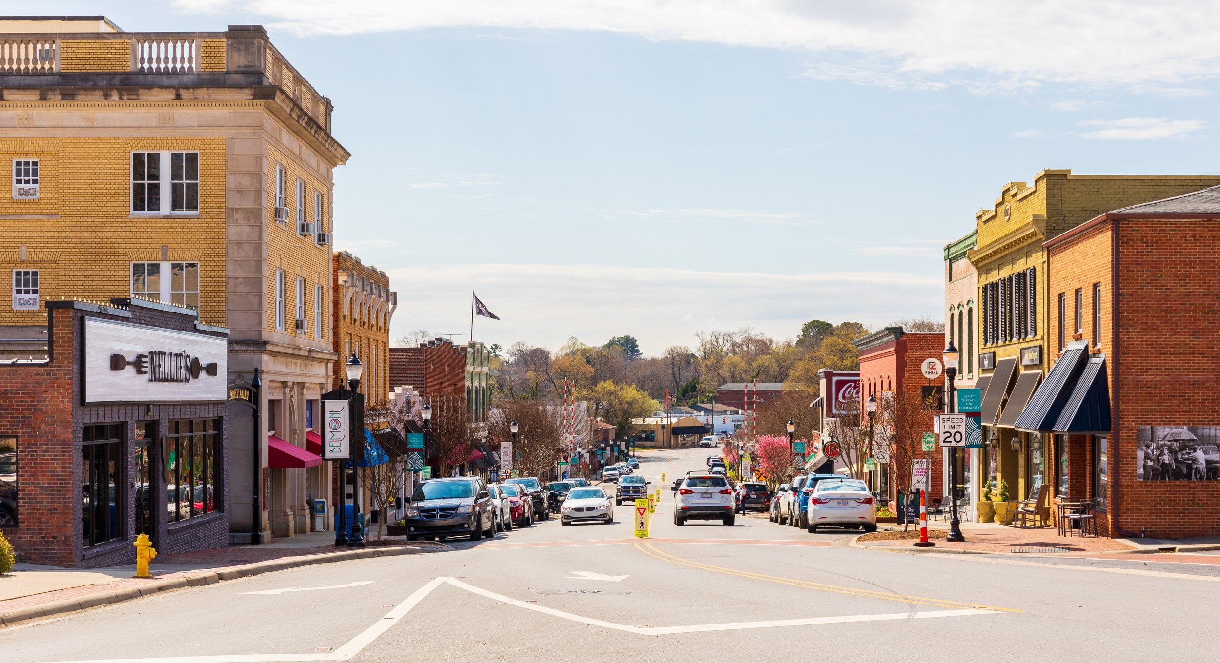 Small downtown street with shops, parked cars on both sides, and trees with pink blossoms. Buildings are brick and some have awnings; the sky is partly cloudy.