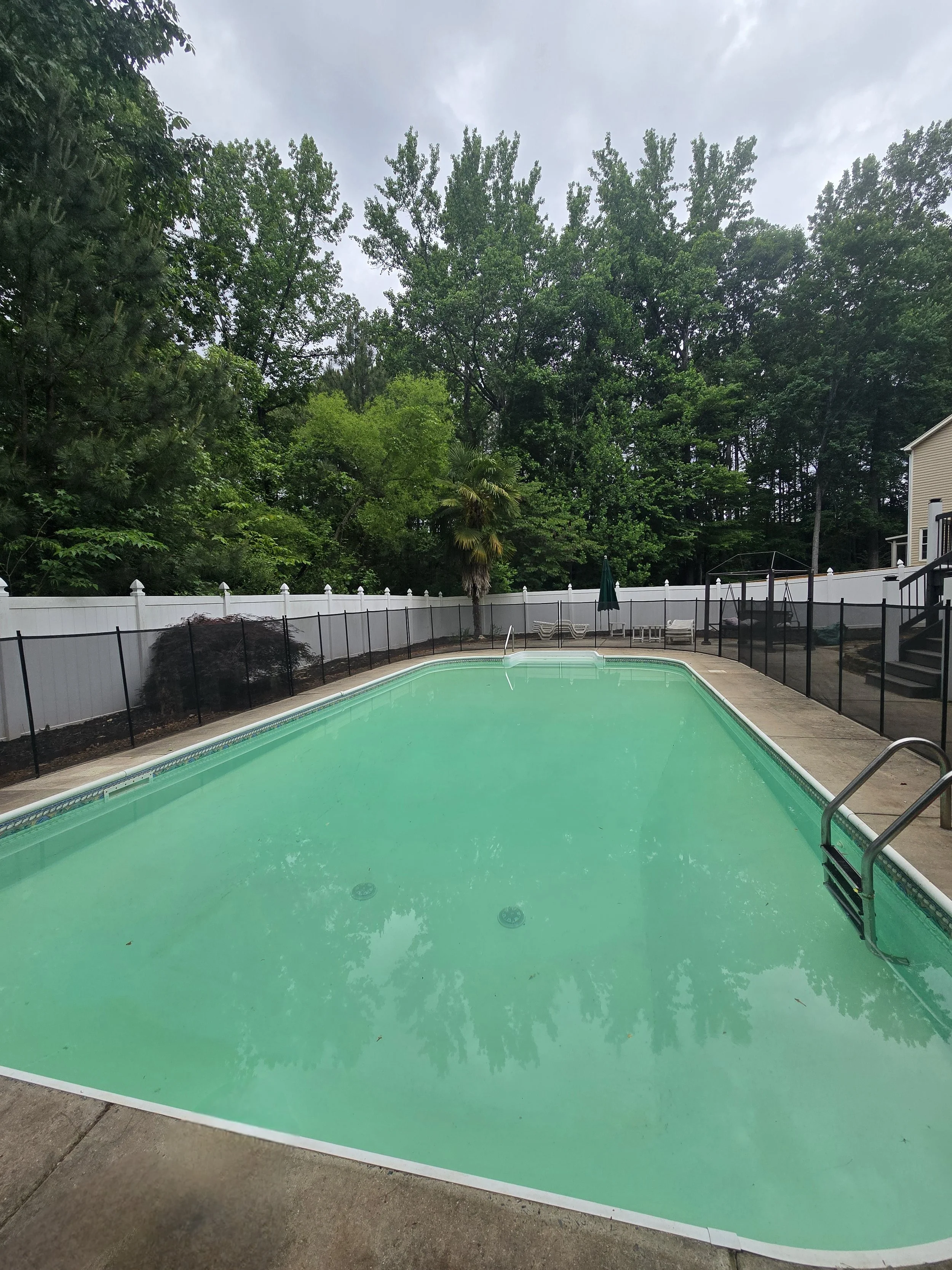 An outdoor swimming pool with clean water, after being serviced by AB Premium Aqua Solutions, surrounded by a white fence. There are lounge chairs and a closed umbrella beside the pool. The background features tall leafy trees and an overcast sky.