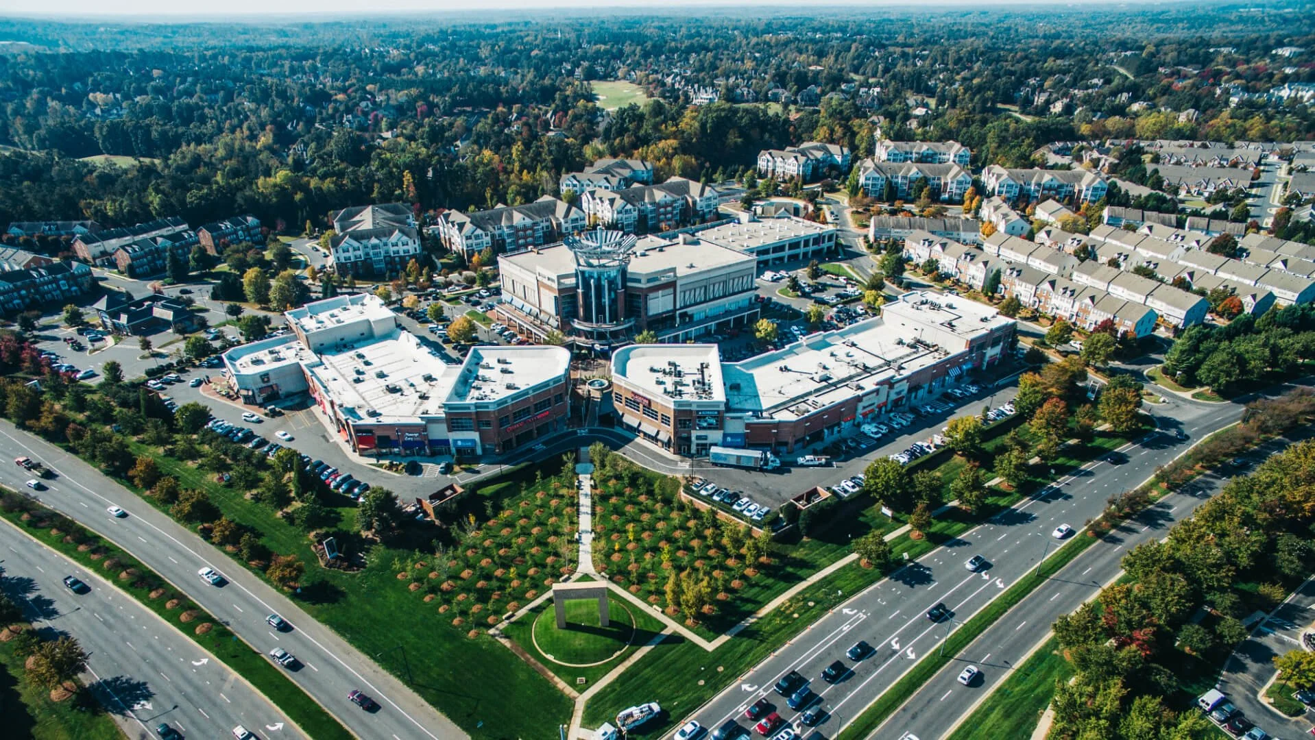 An aerial view of the Ballantyne, NC shopping mall surrounded by parking lots, roads, and green spaces with trees and a park in the foreground, residential houses in the background, and wooded areas further away.