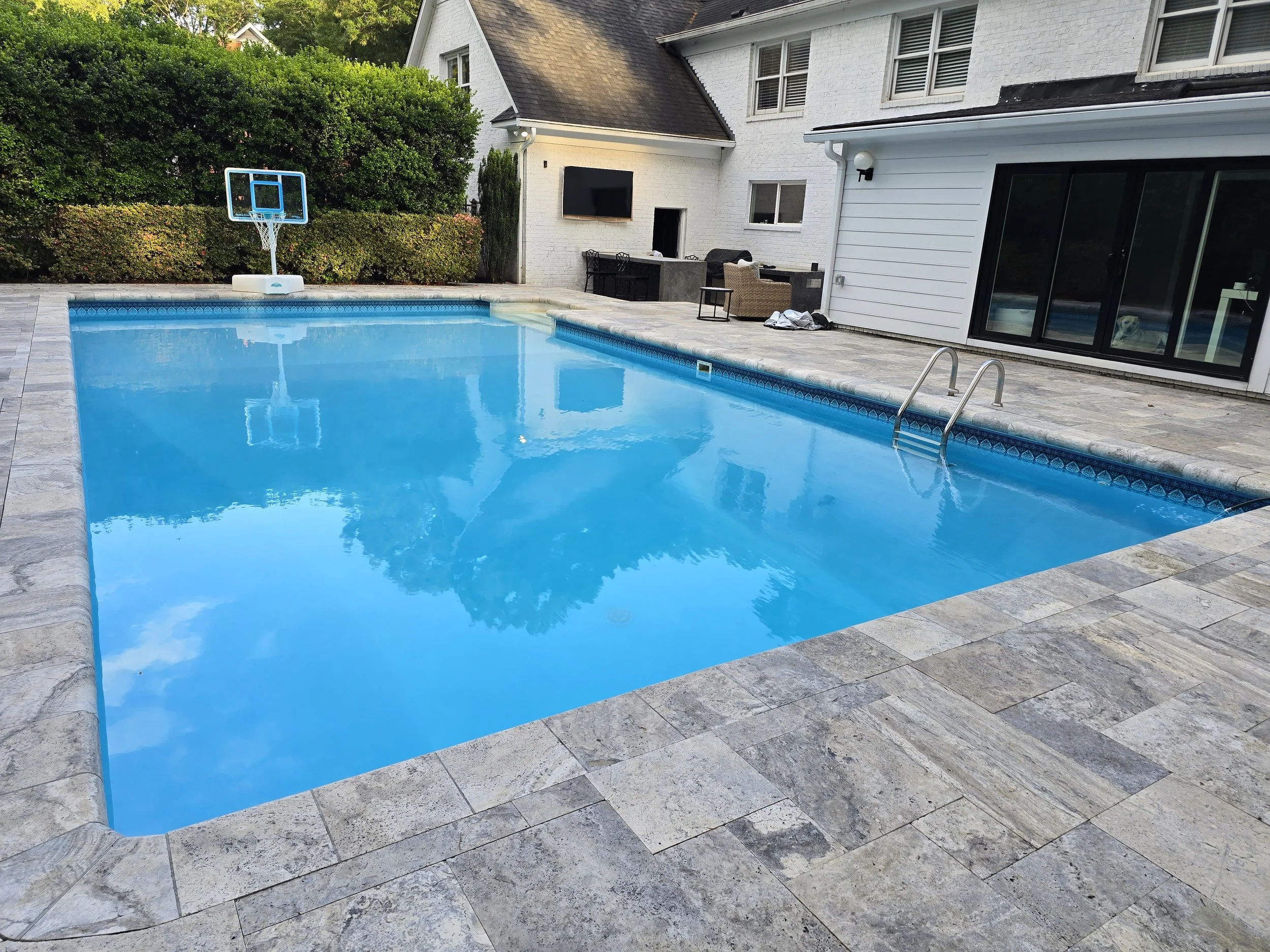 A backyard swimming pool serviced by AB Premium Aqua Solutions, with a blue basketball hoop at one end and the surrounding paving gray stone, with backyard furniture and greenery visible near the white house.