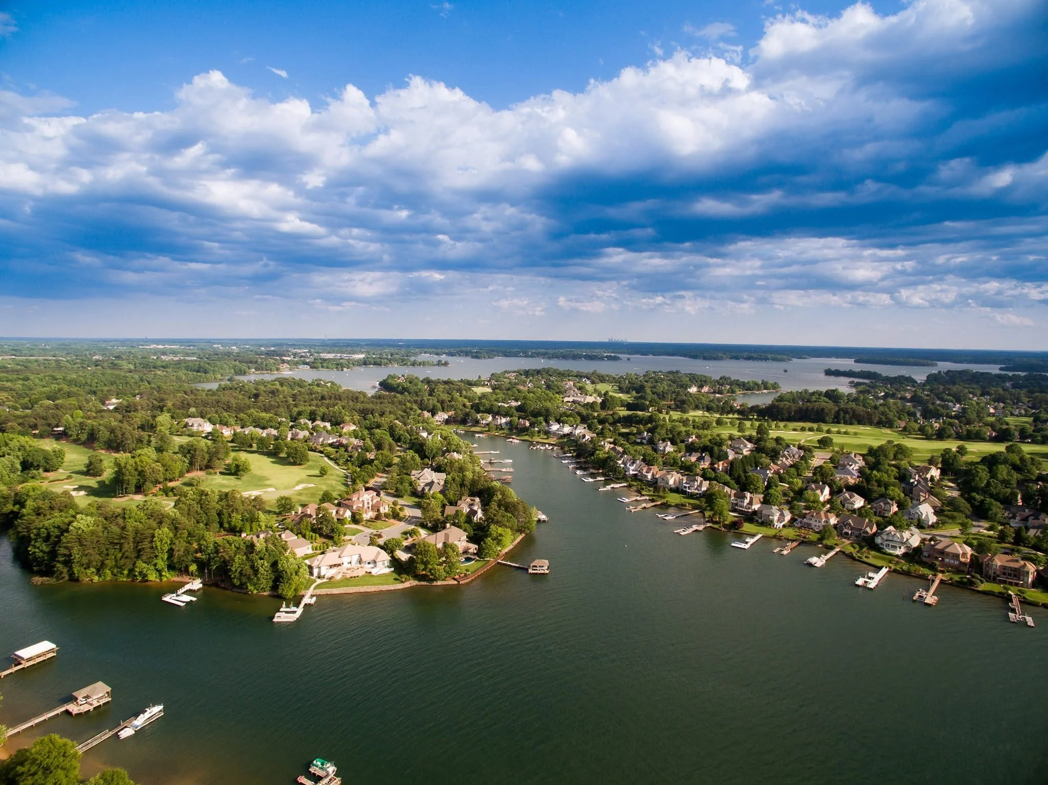 Aerial view of a lake with surrounding residential houses, boats at docks, and lush green trees under a partly cloudy sky.