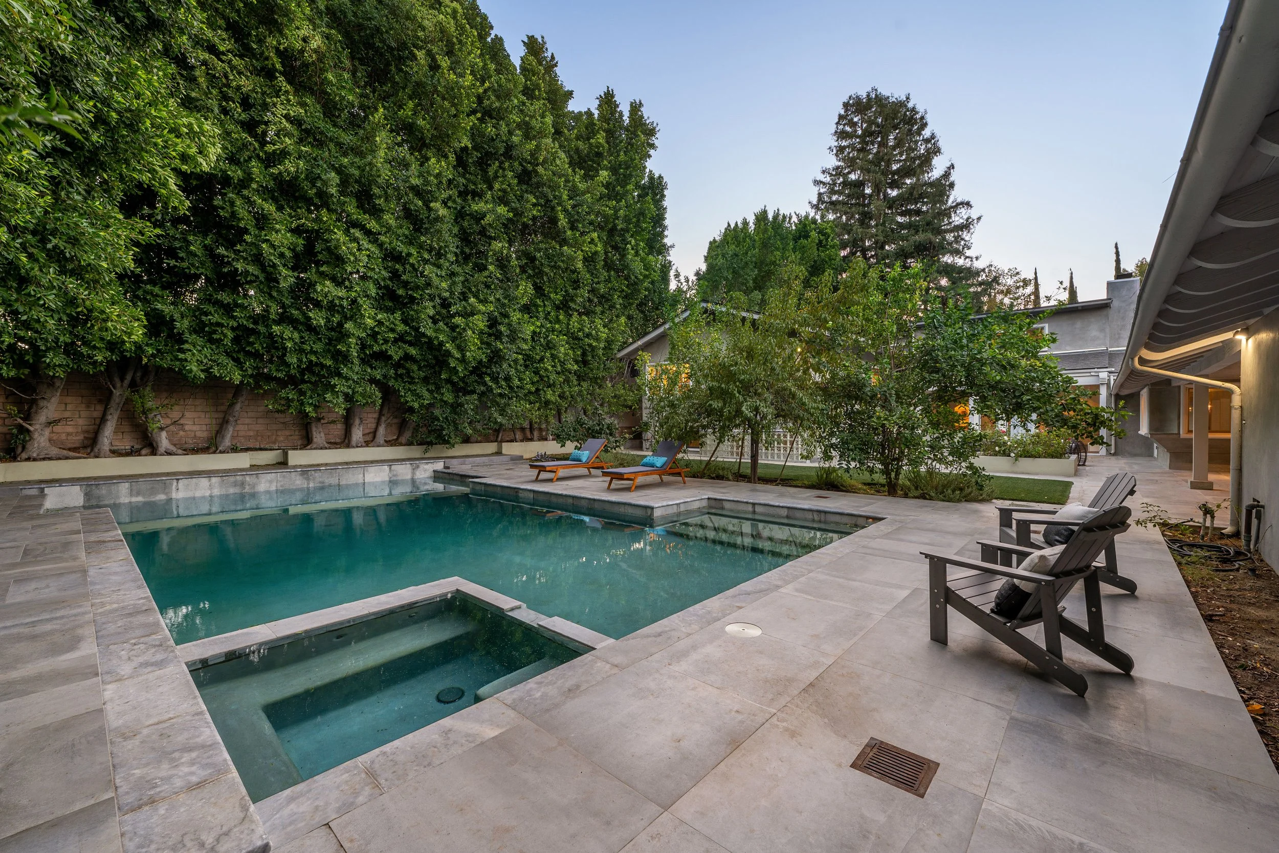 A backyard with a swimming pool serviced by AB Premium Aqua Solutions, lounge chairs, trees, and a house on a clear evening.