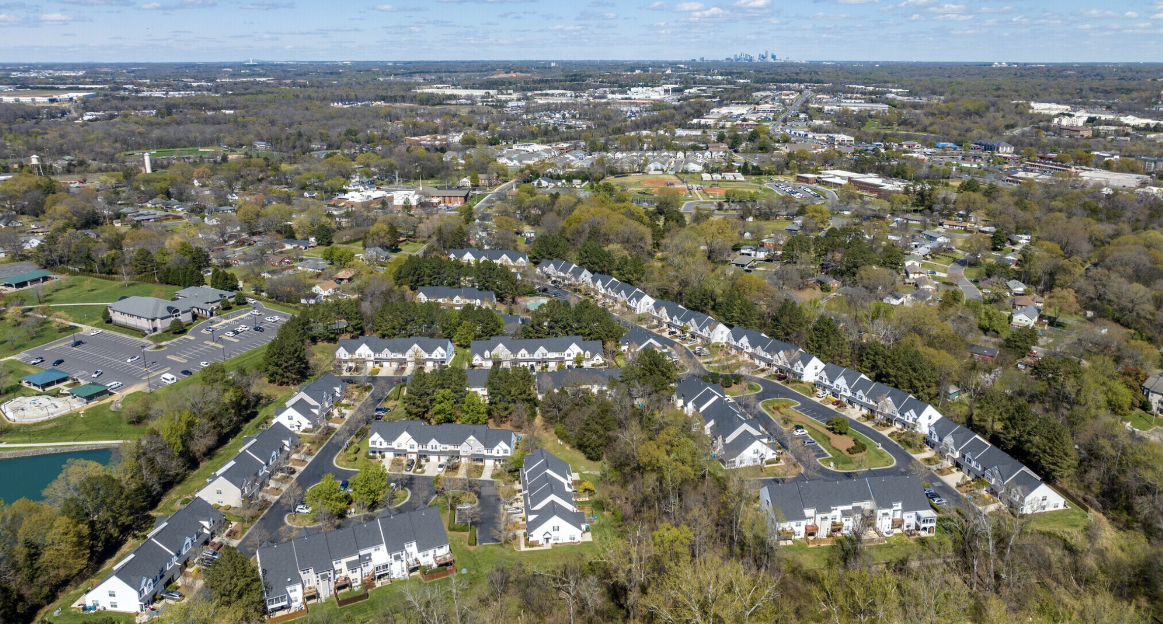 Aerial view of a suburban neighborhood with rows of white houses with dark roofs, surrounded by trees and parking lots. In the distance, a city skyline is visible under a partly cloudy sky.