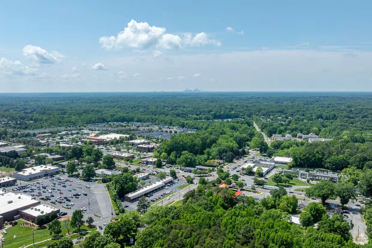 Aerial view of a suburban area with a large green forest in the background, a commercial complex, roads, and parking lots, with a city skyline faintly visible on the horizon under a partly cloudy sky.