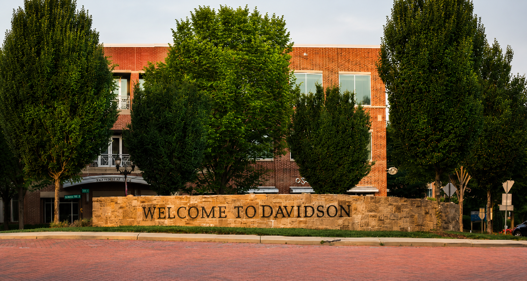 A brick building behind a stone wall with a sign that reads 'WELCOME TO DAVIDSON' in front of a row of trees and a brick sidewalk.