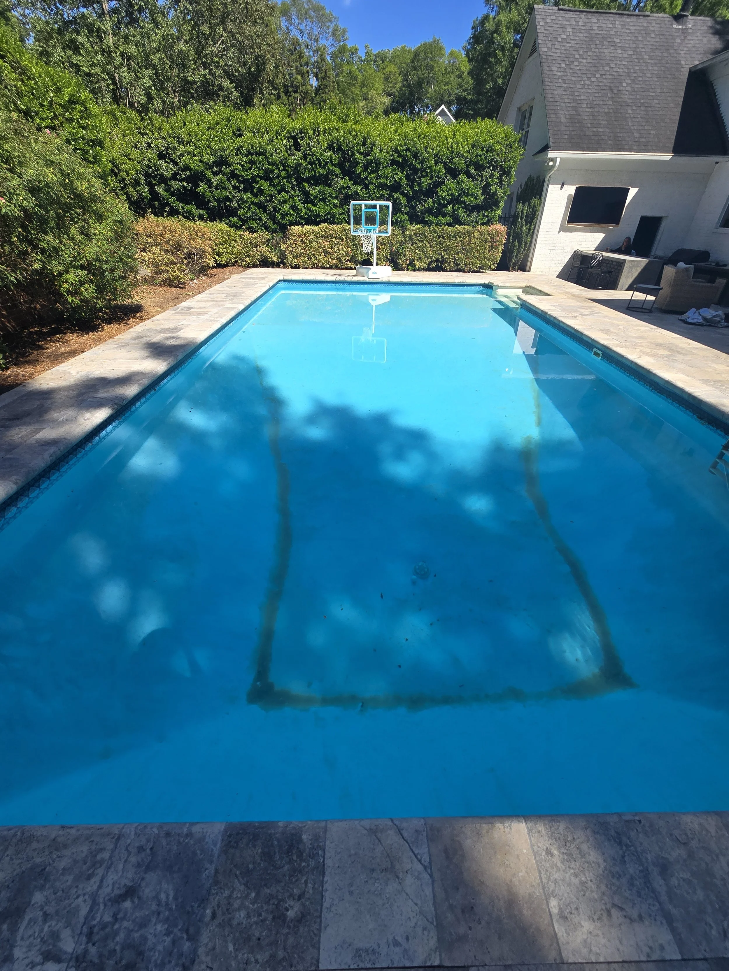 A backyard swimming pool during treatment by AB Premium Aqua Solutions, with a blue basketball hoop mounted at the far end, surrounded by a stone deck, with green bushes and tall trees in the background, and part of a white house.