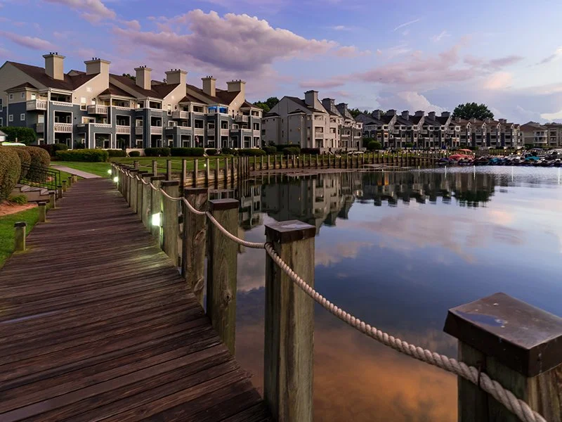 A wooden dock with rope railing runs along the edge of a calm lake, with modern multi-story apartment buildings in the background under a sky with scattered clouds during sunset.