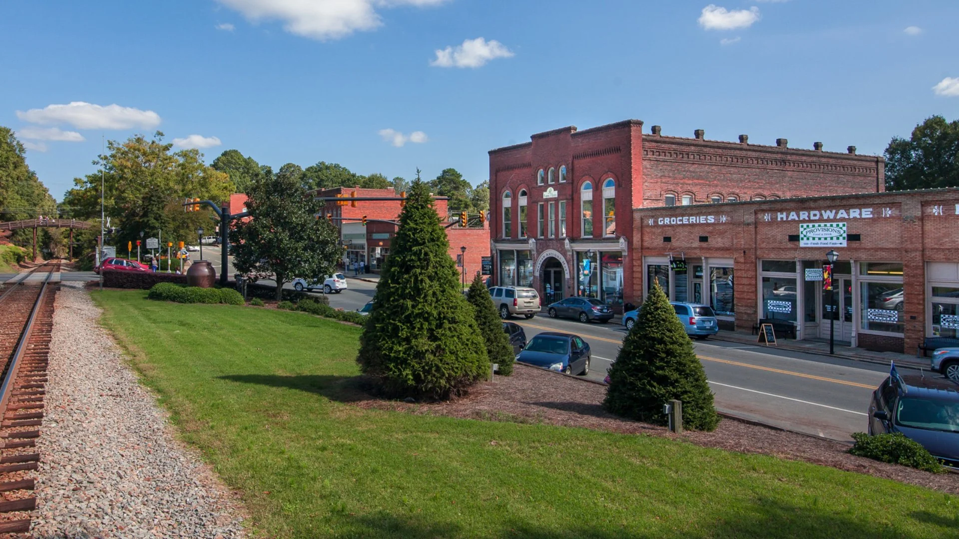 A small downtown area with brick buildings, a street, parked cars, green trees, and a railroad track in the foreground.