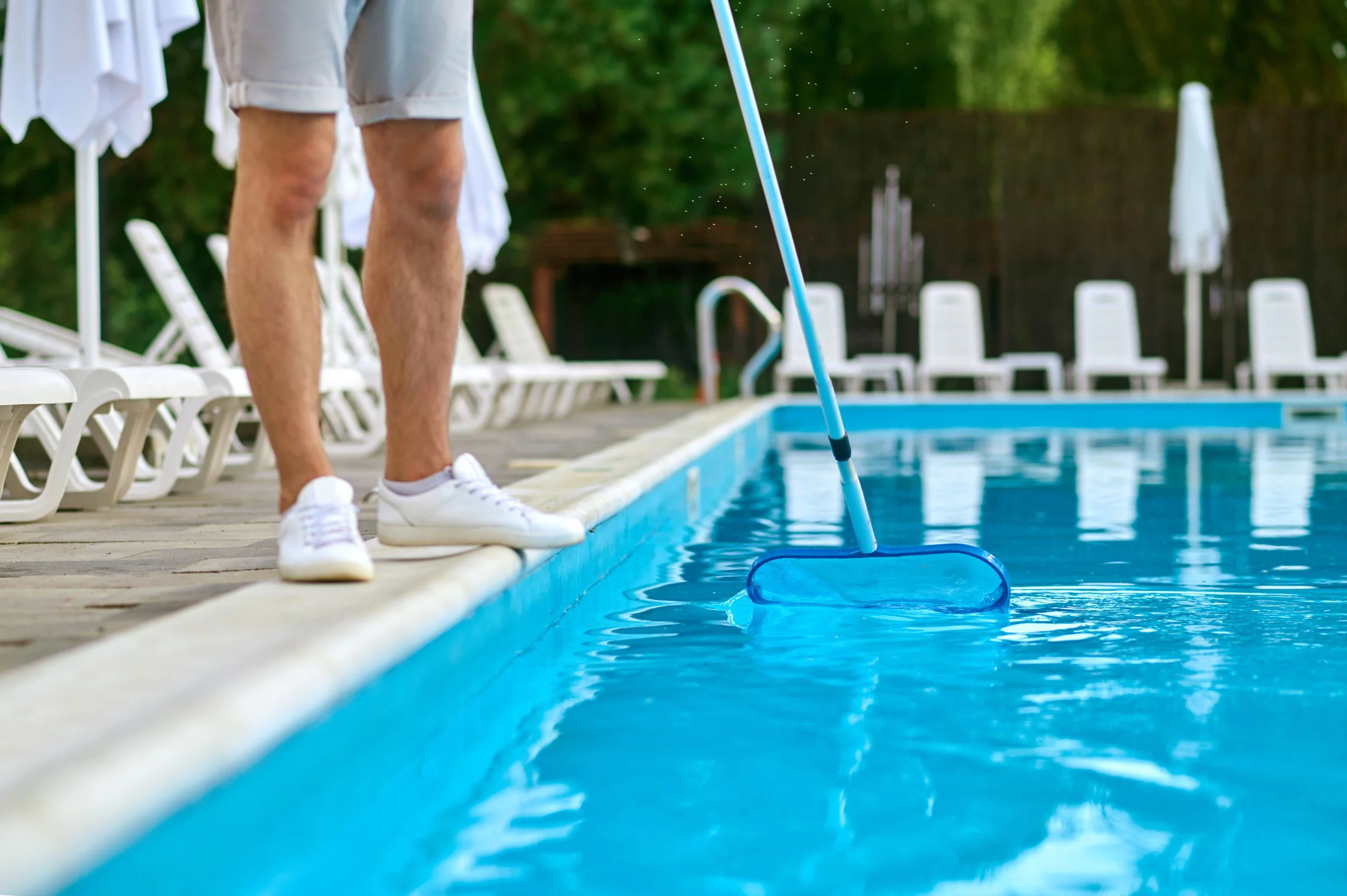 AB Premium Aqua Solutions staff in white shorts and sneakers cleaning the pool with a blue pool skimmer, poolside chairs and umbrellas in the background.