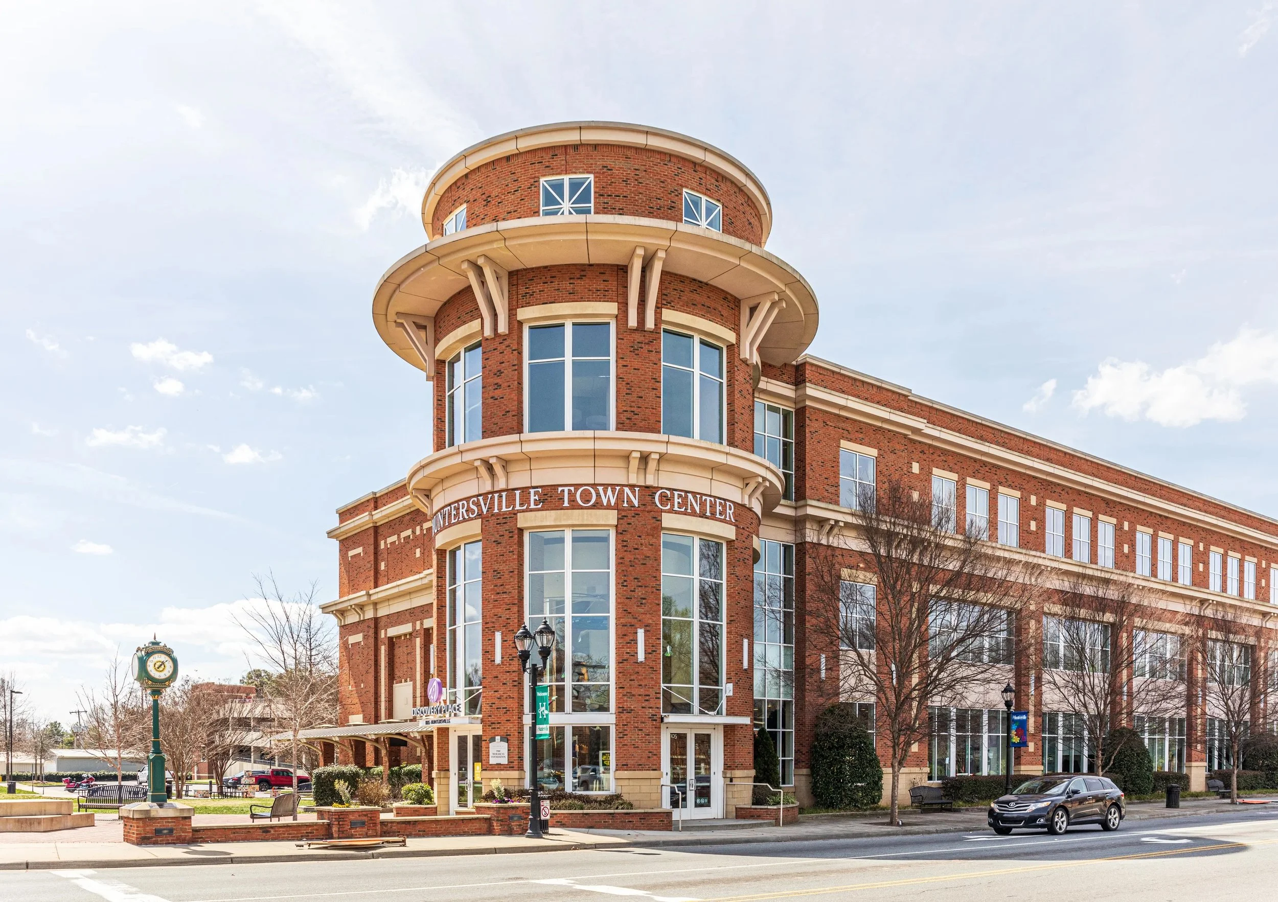 A red brick building with rounded towers at the top, large windows, and a sign that reads 'Interstate Town Center.' There is a sidewalk, benches, and a lamp post in front of the building, with a street with cars passing by and a clock tower visible in the background.