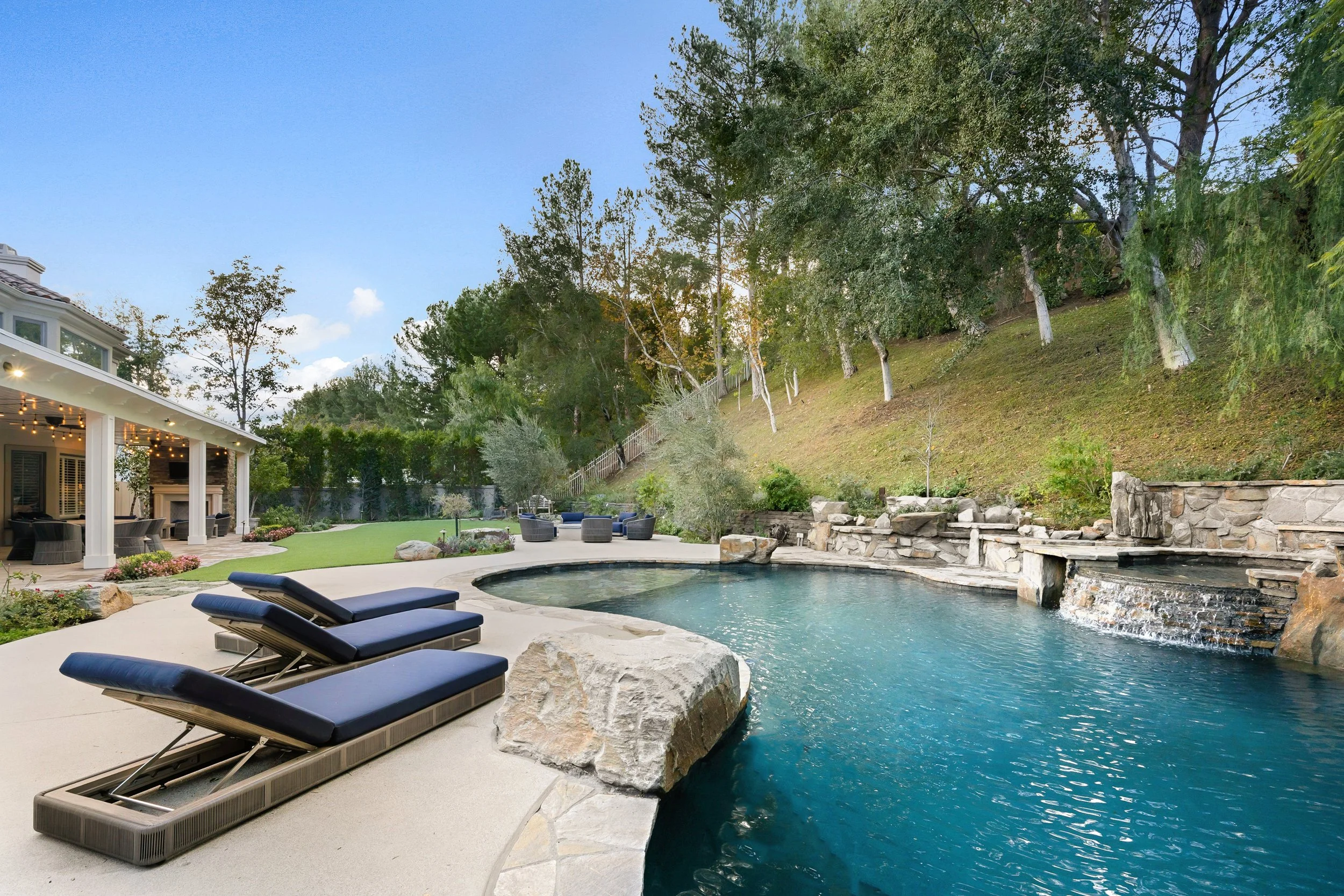 A backyard swimming pool serviced by AB Premium Aqua Solutions with lounge chairs, a covered patio with lights, and a hillside with trees and a fence.