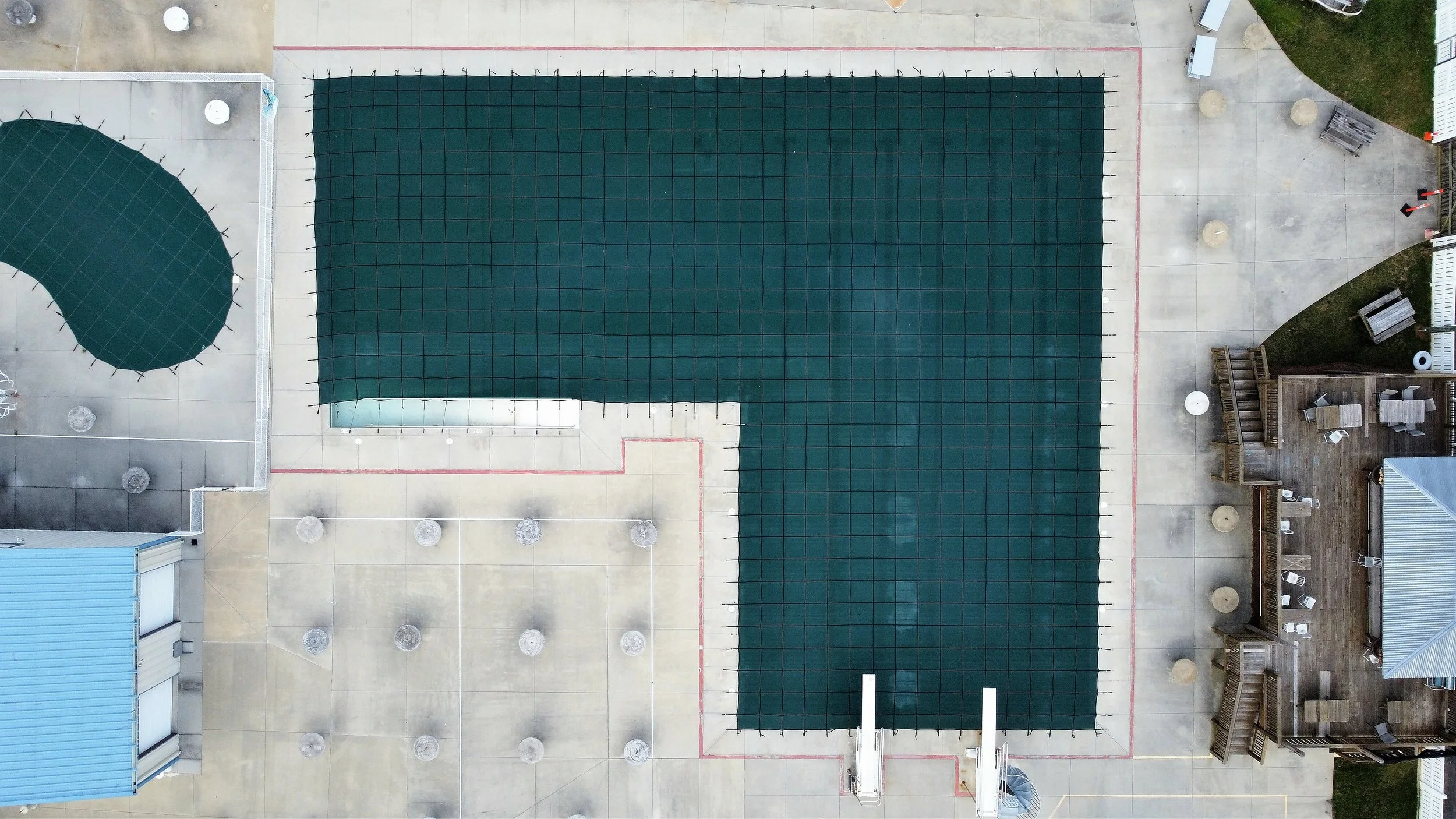 An aerial view of an outdoor pool serviced by AB Premium Aqua Solutions with a large rectangular green cover and a smaller rounded green cover, with surrounding concrete and wooden deck areas that have seating and umbrellas.