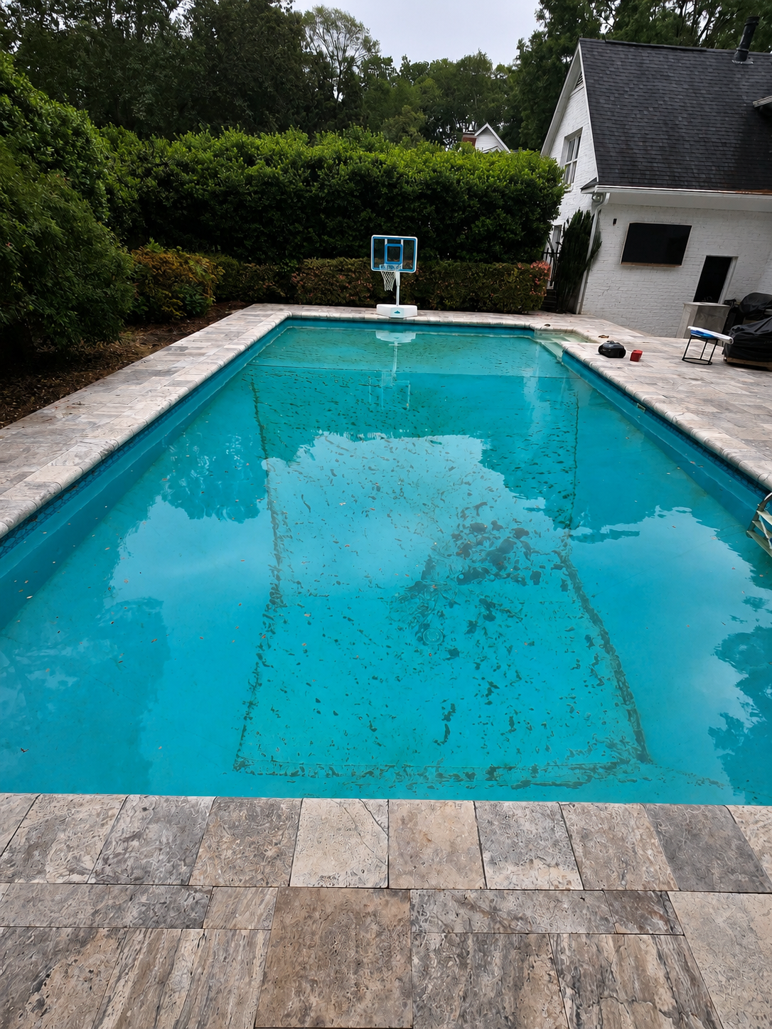 A backyard swimming pool during treatment by AB Premium Aqua Solutions with leaves floating on the water, surrounded by stone tiles, with a small basketball hoop at the end, bushes, and a house in the background.