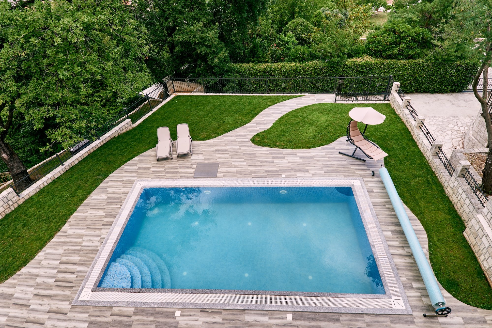 A backyard with a rectangular swimming pool serviced by AB Premium Aqua Solutions, a green lawn, lounge chairs, a patio umbrella, and surrounding trees and bushes.