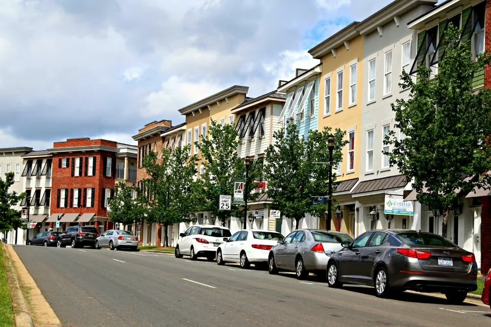 A street lined with multi-story residential buildings with retail shops on the ground floor. Several cars are parked along the curb, and green trees are interspersed between the buildings. The sky is partly cloudy.