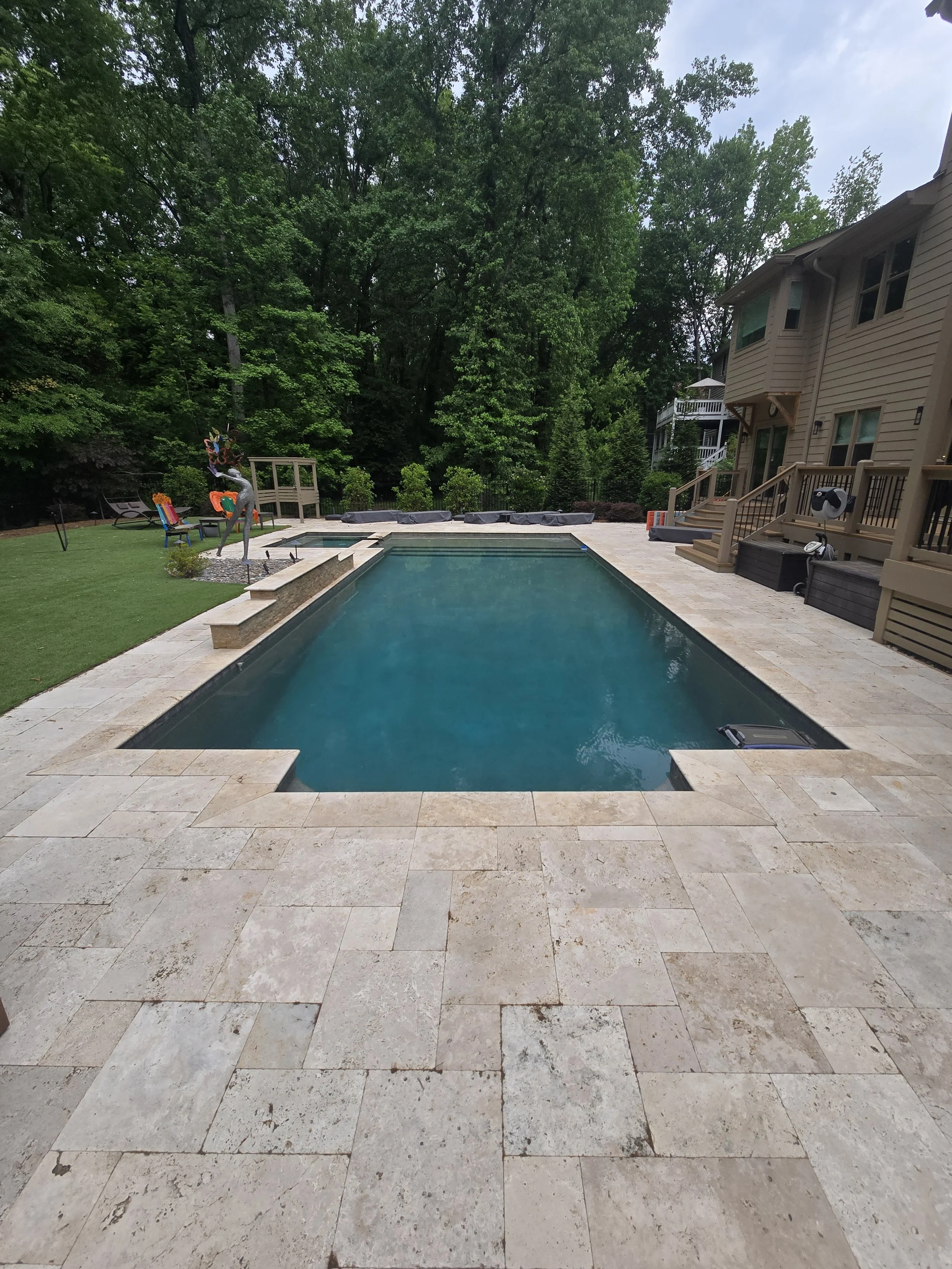 A backyard with a rectangular swimming pool serviced by AB Premium Aqua Solutions, with a stone patio, green grass, garden statues, trees, and a wooden house.