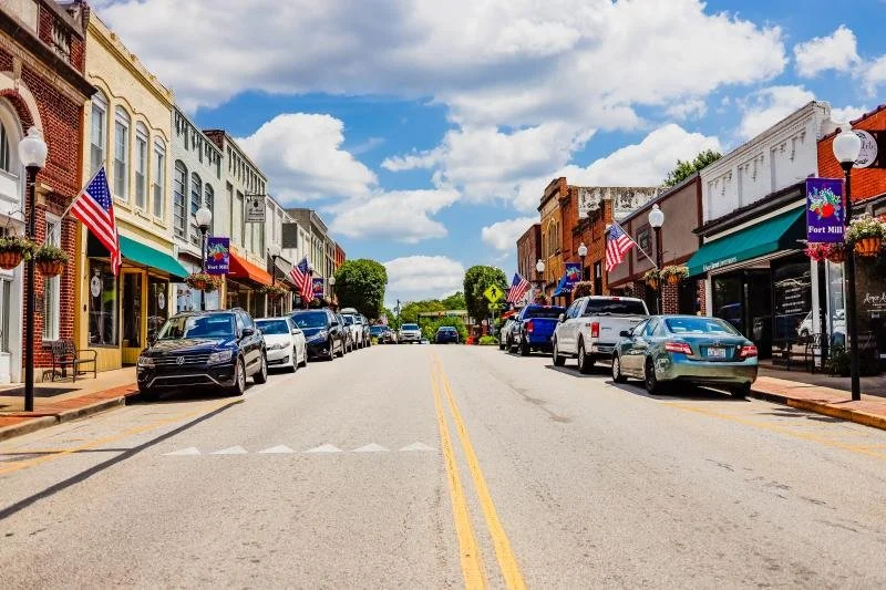 A sunny street in a small American town with cars parked along both sides, American flags hanging from lampposts, and small shops with colorful awnings.