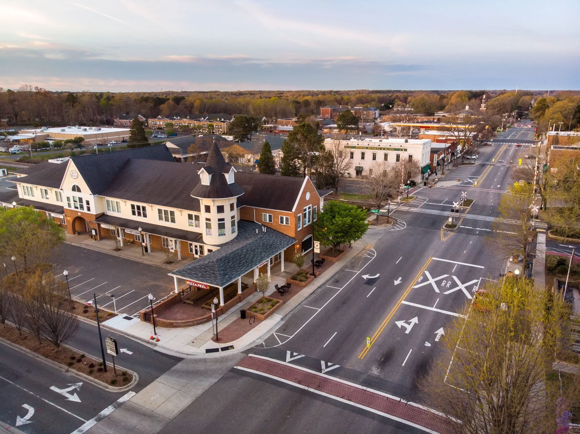 An aerial view of a small town with commercial buildings, parking lots, and streets, surrounded by trees and rolling hills.