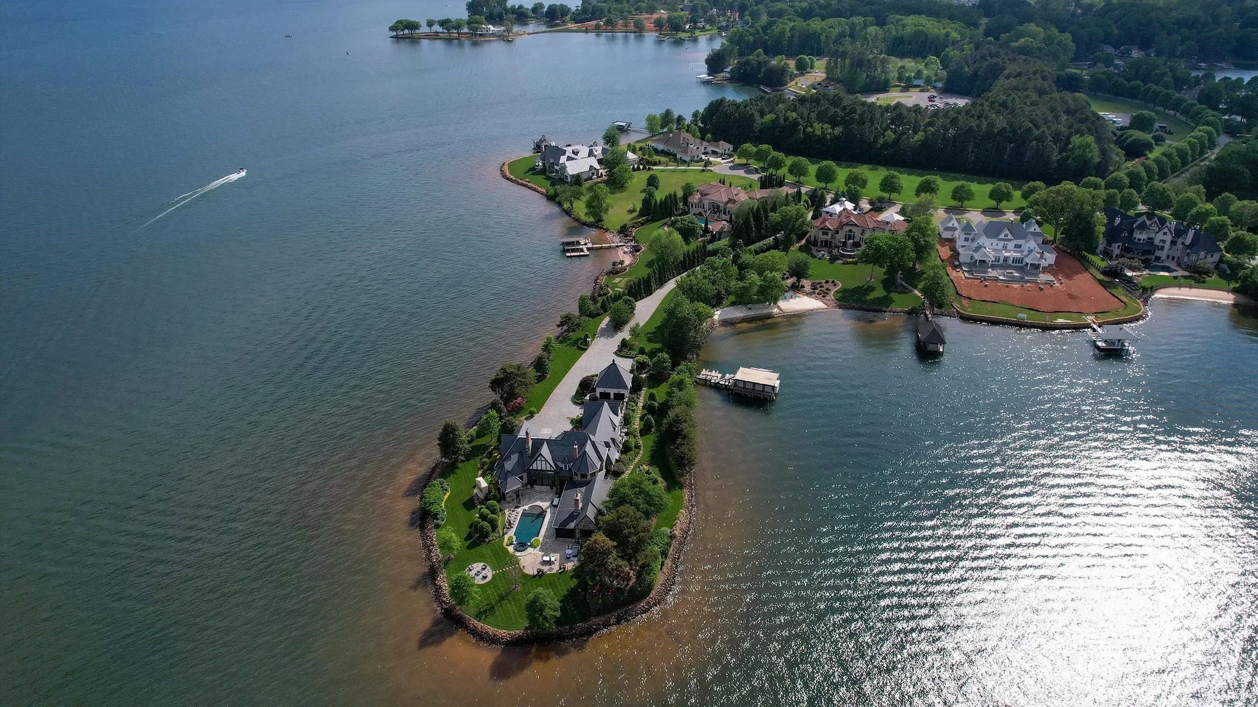 Aerial view of a lakeside neighborhood with large houses, green lawns, trees, and docks along the water. A boat is seen traveling across the lake, creating a wake.