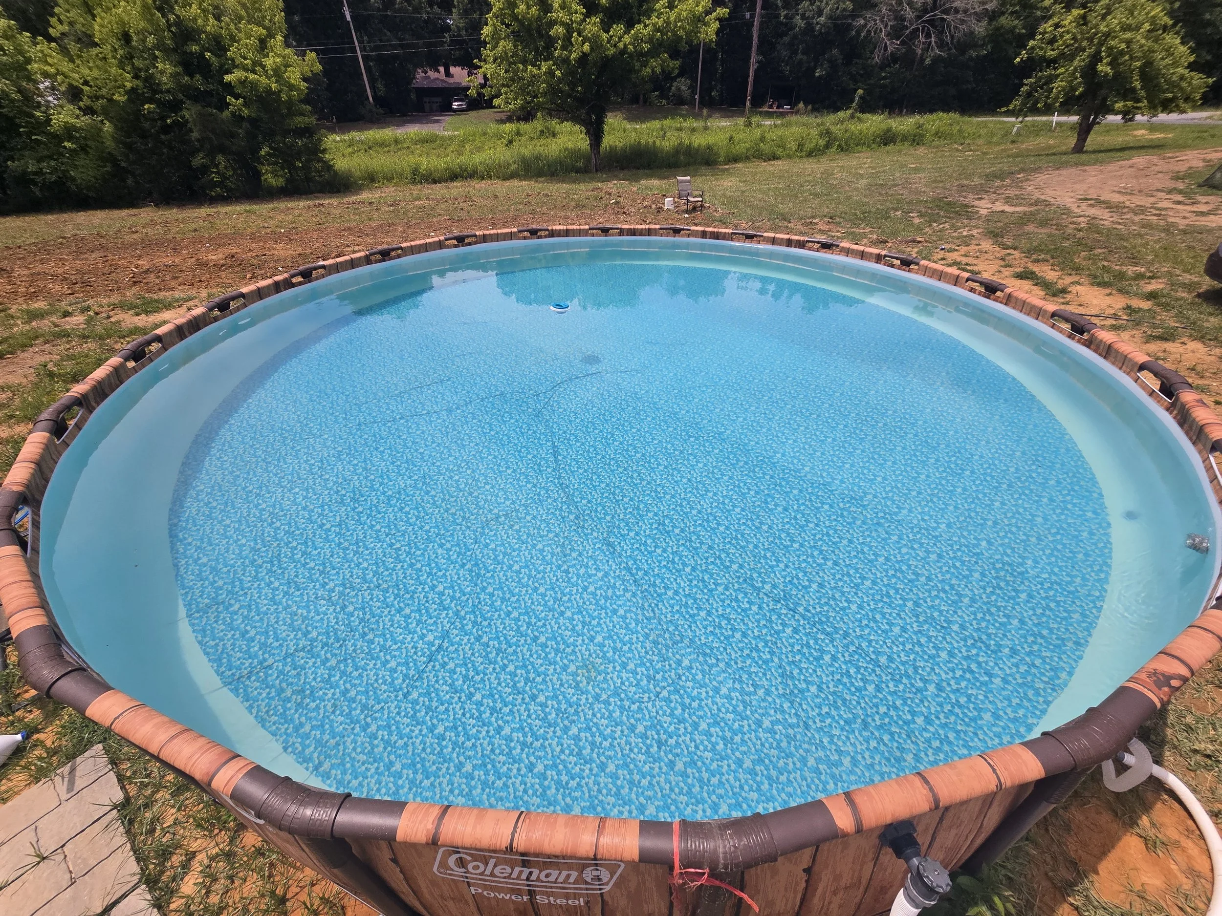 A round above-ground swimming pool filled with blue water, after being serviced by AB Premium Aqua Solutions, surrounded by a brick and plastic border, set on a patchy grassy and dirt yard with trees and a house in the background.