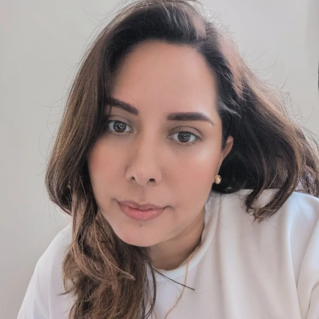 A woman with medium-length brown hair, wearing a white top and gold earrings, looking into the camera with a neutral expression.