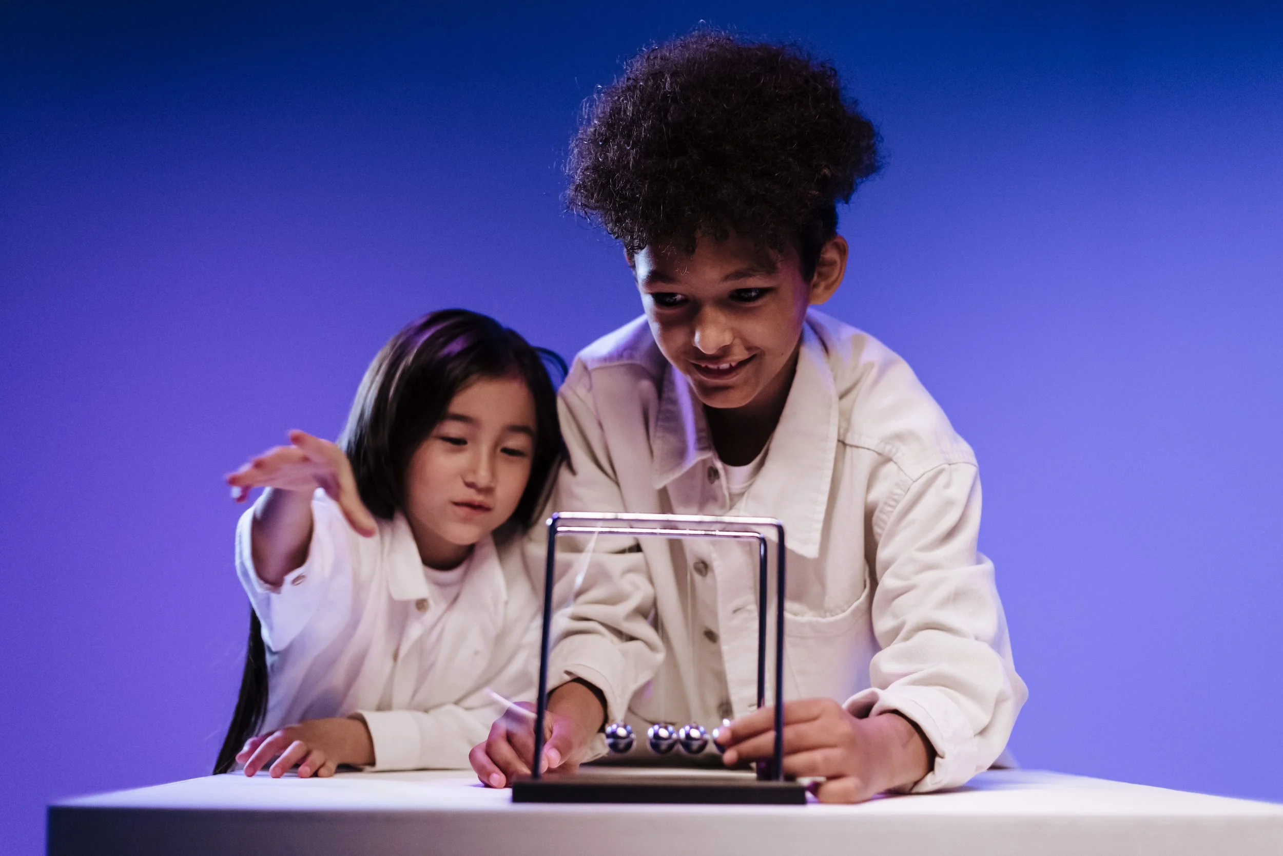 Two children, a girl with long dark hair and a boy with curly black hair, are playing a game with a small spherical object in front of a purple background.