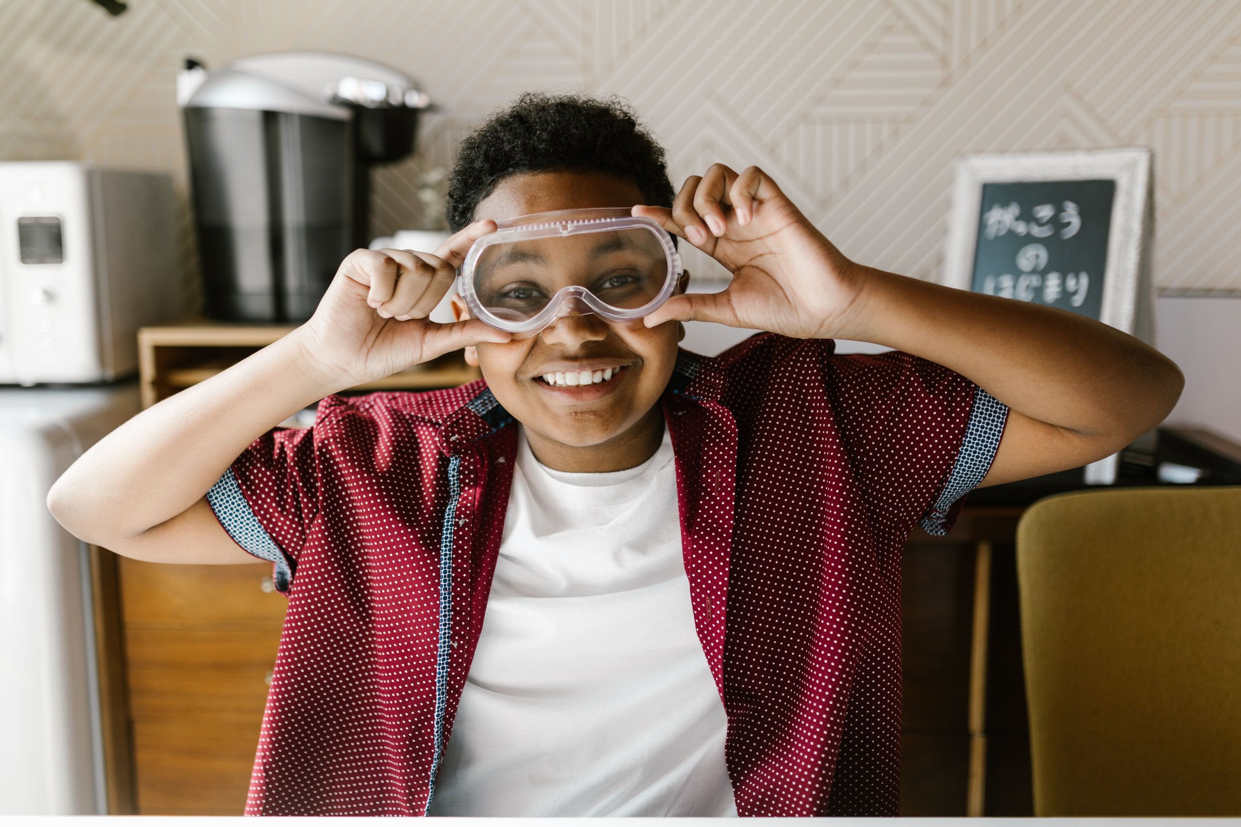 Smiling child in red shirt holding protective goggles up to their eyes.