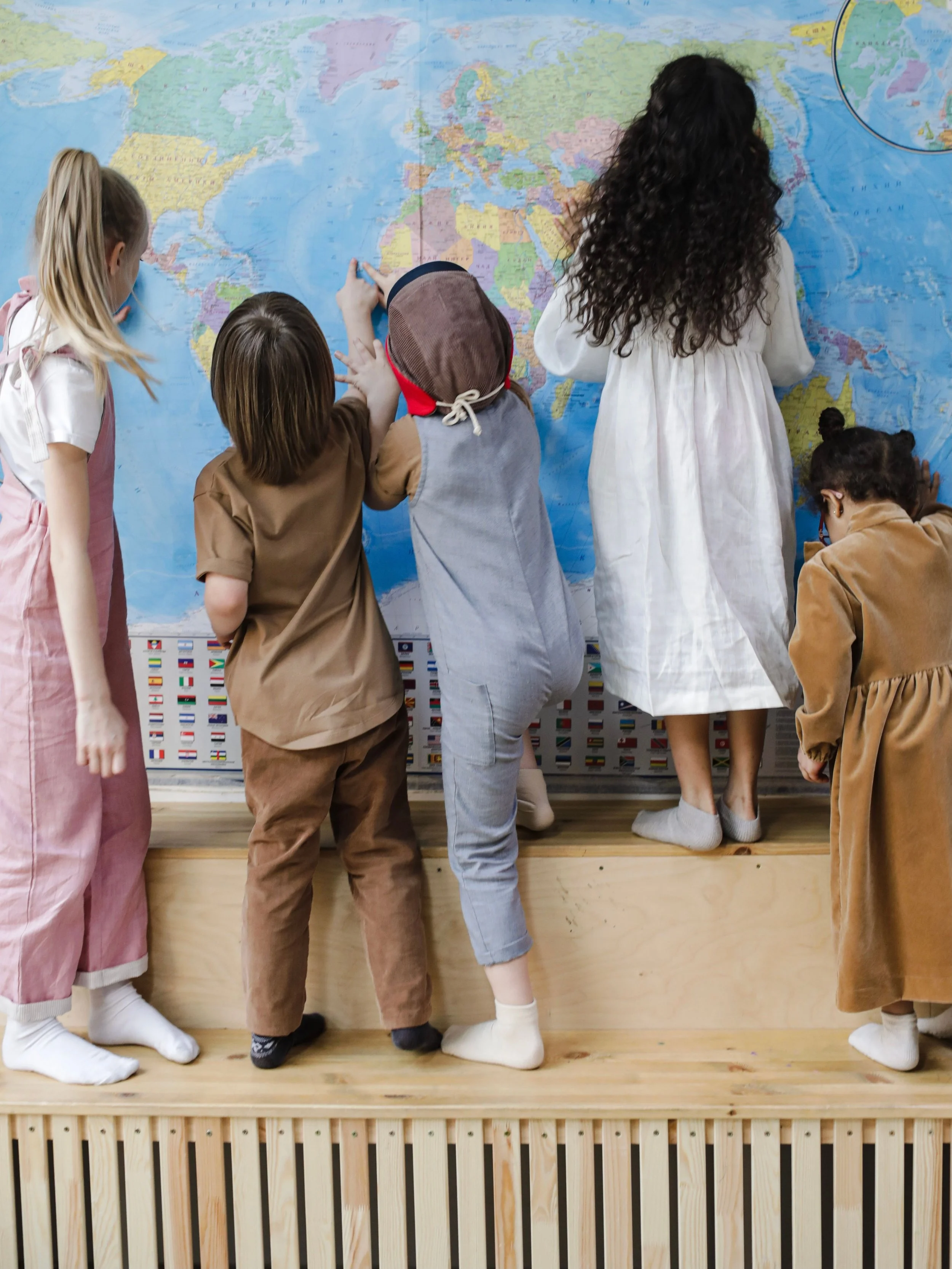 Children dressed in vintage clothing, some wearing head coverings, looking at and pointing to a large world map on a classroom wall.