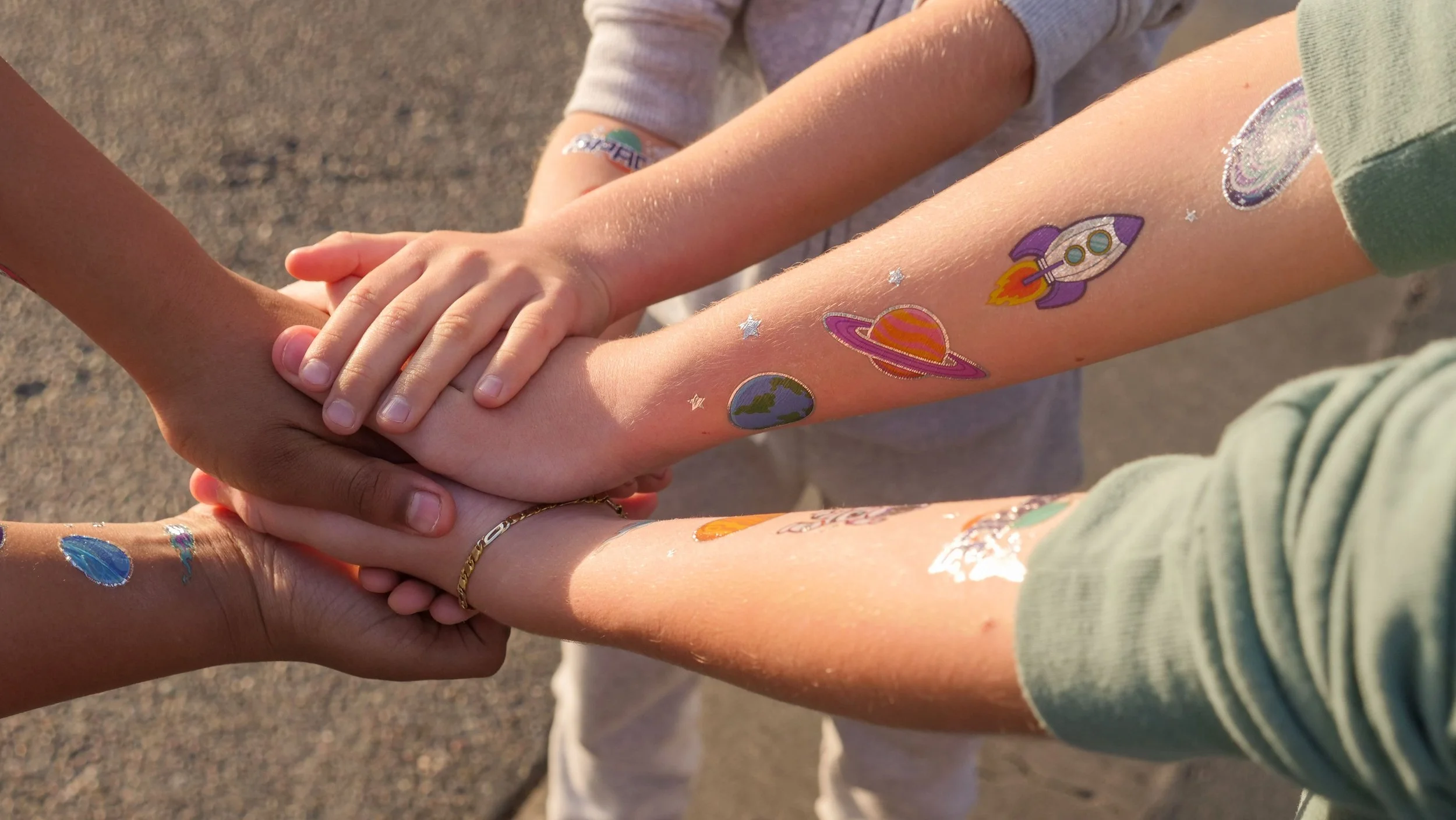 Four people hold hands in a circle, showing colorful space-themed temporary tattoos on their arms and legs.