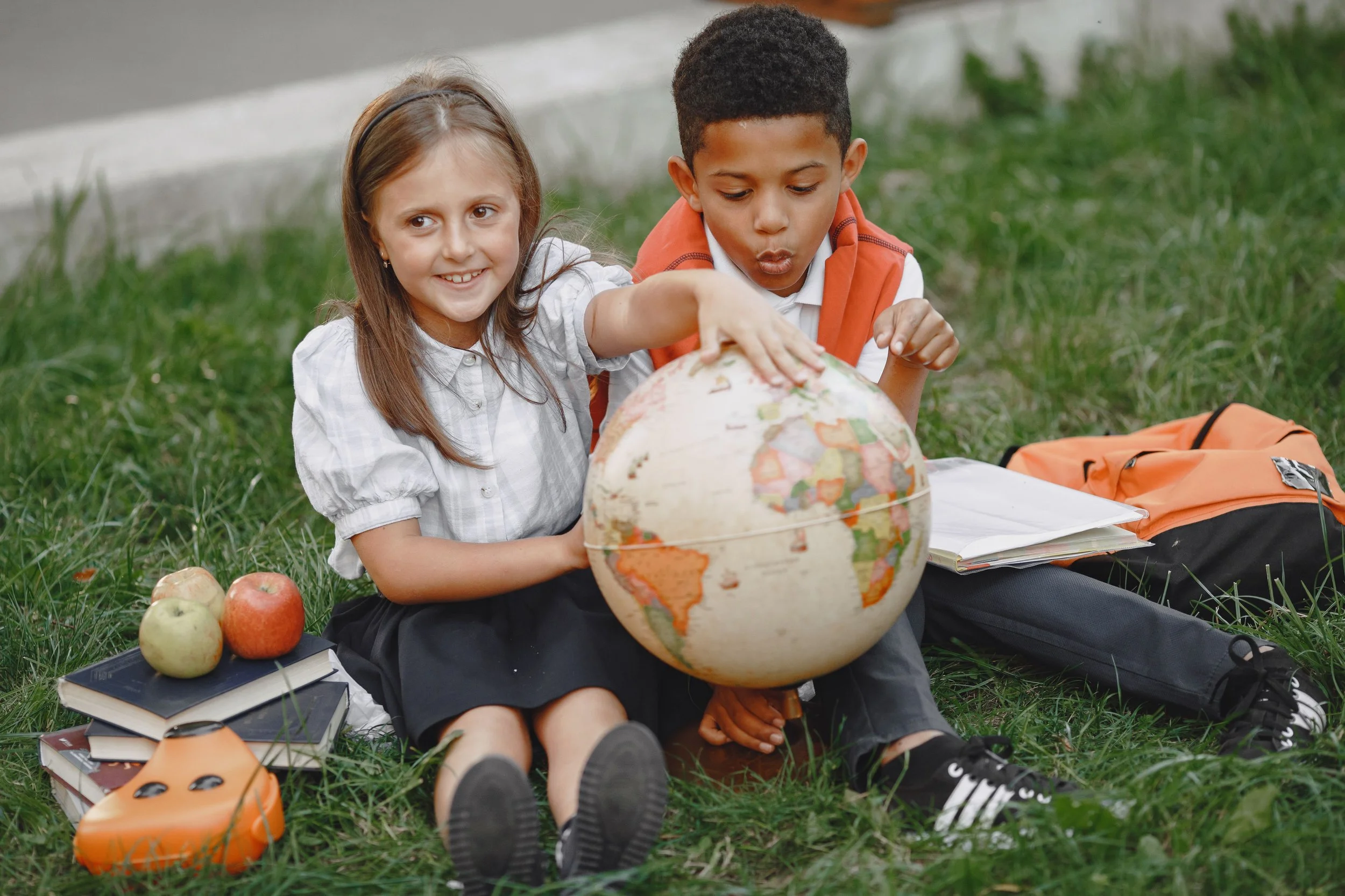 Two children, a girl and a boy, sitting on grass outdoors with a globe in front of them. The girl is smiling and pointing at the globe while the boy is blowing on it. Apples, books, and a backpack are nearby.