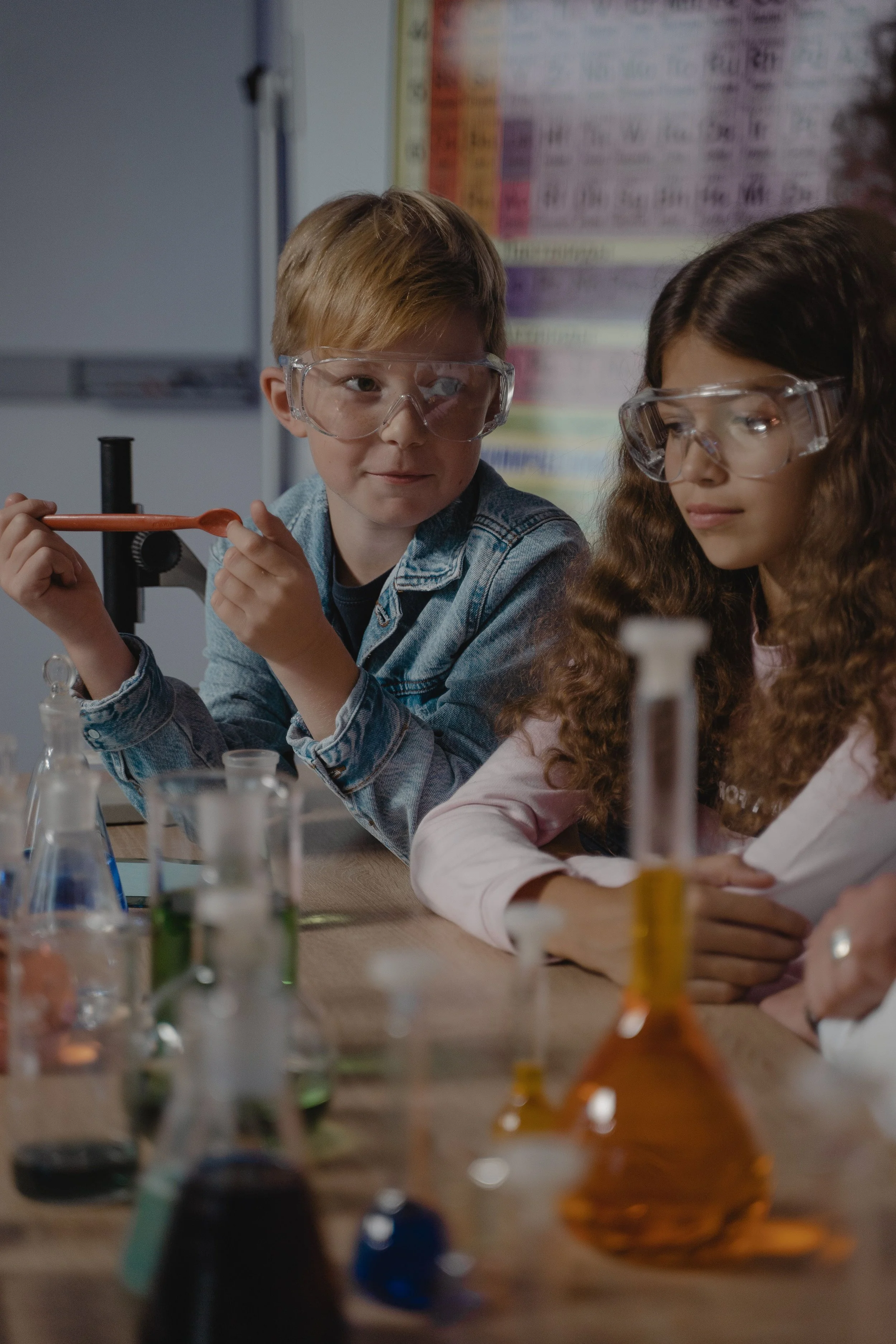 Two children, a boy and a girl, wearing safety goggles in a science classroom, working with various laboratory glassware and chemicals on a table.