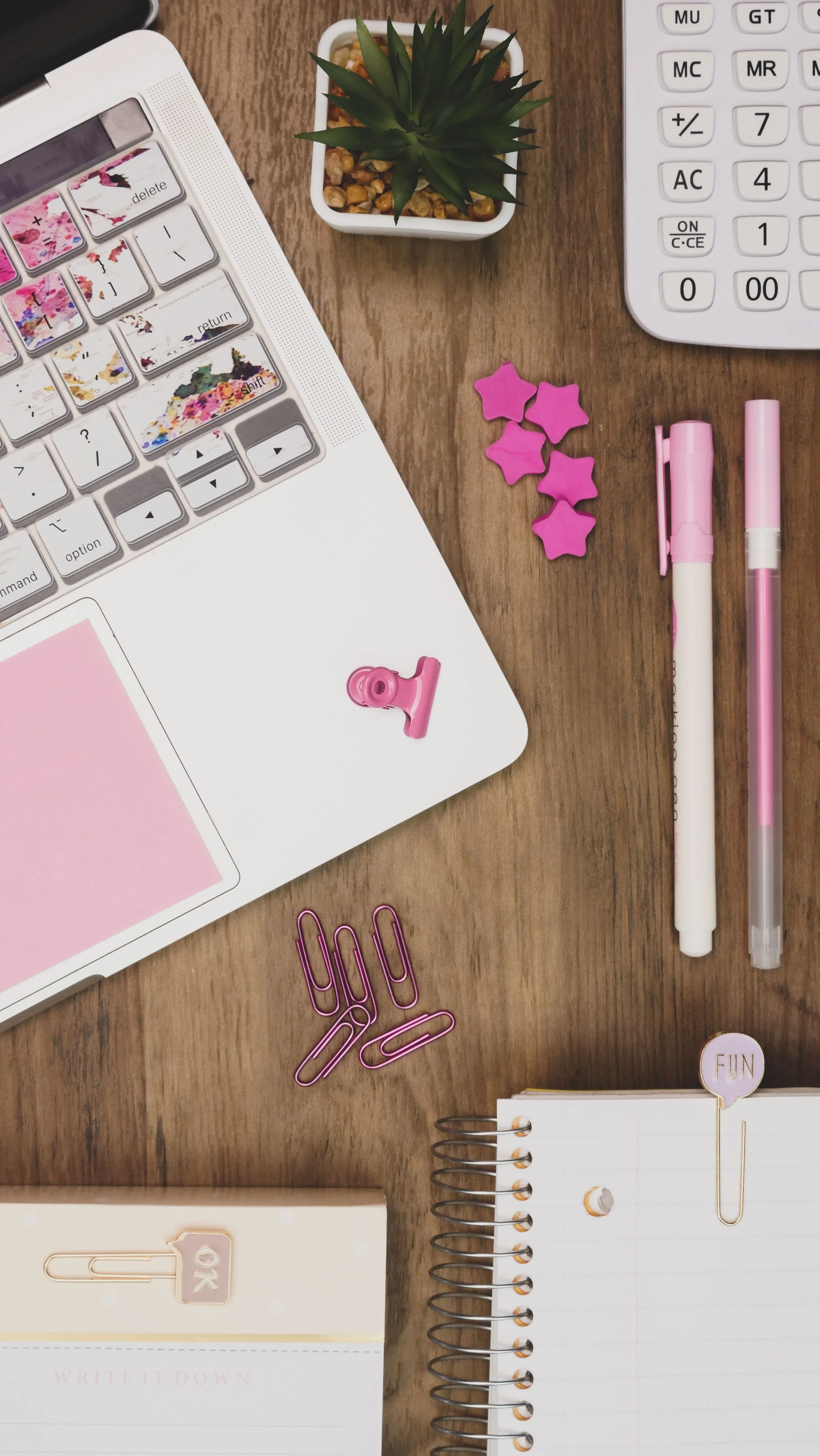 Flat lay of a workspace with a laptop, a small potted succulent, a white calculator, pink star-shaped clips, pink and white markers, pink paper clips, a beige Notepad with a sticker that says 'XOXO', and a spiral-bound notebook with a speech bubble that says 'FUN' on the cover.