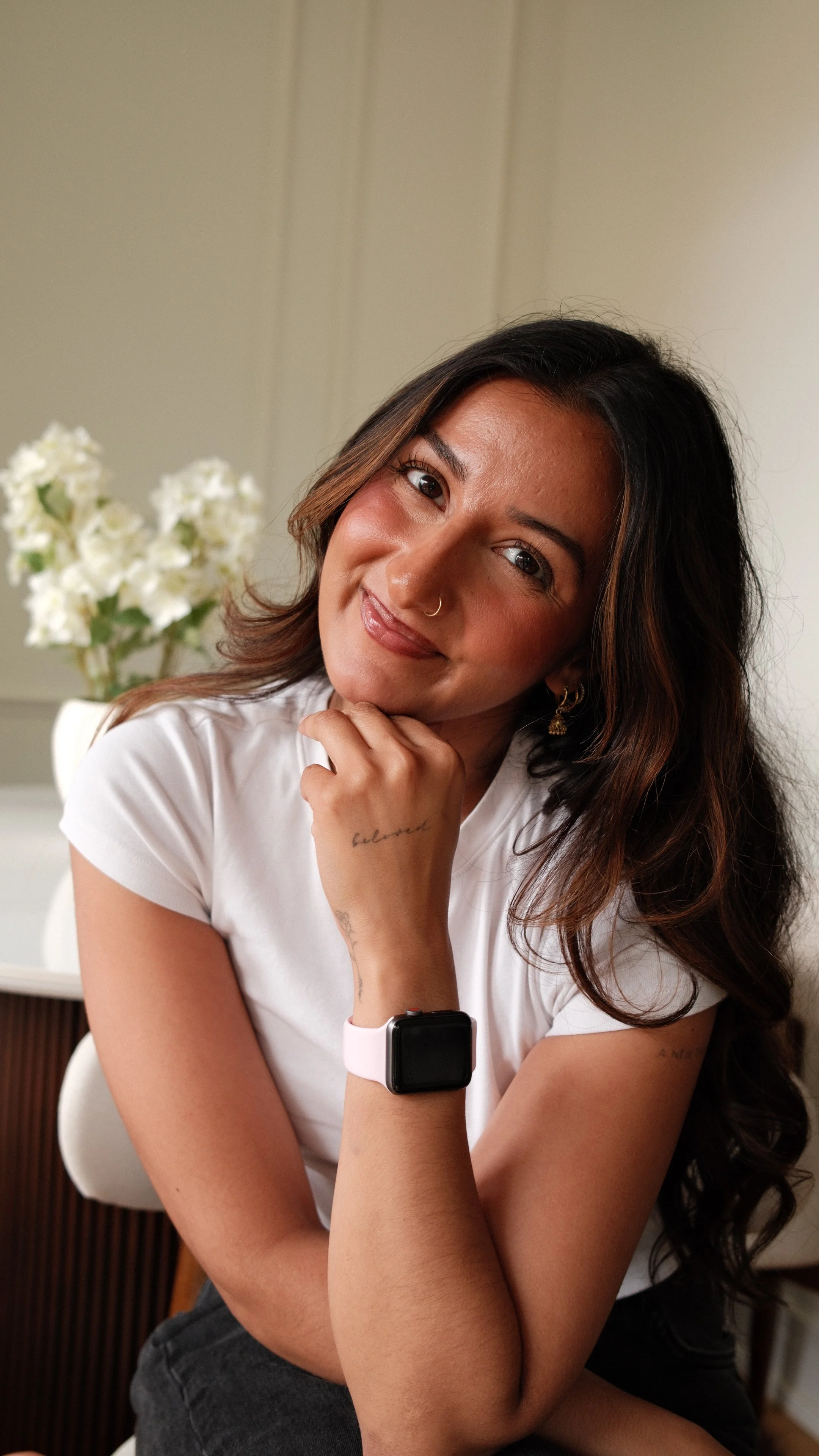 A woman with dark hair, wearing a white shirt and a smartwatch, sitting indoors with her hand resting on her chin, smiling softly at the camera.