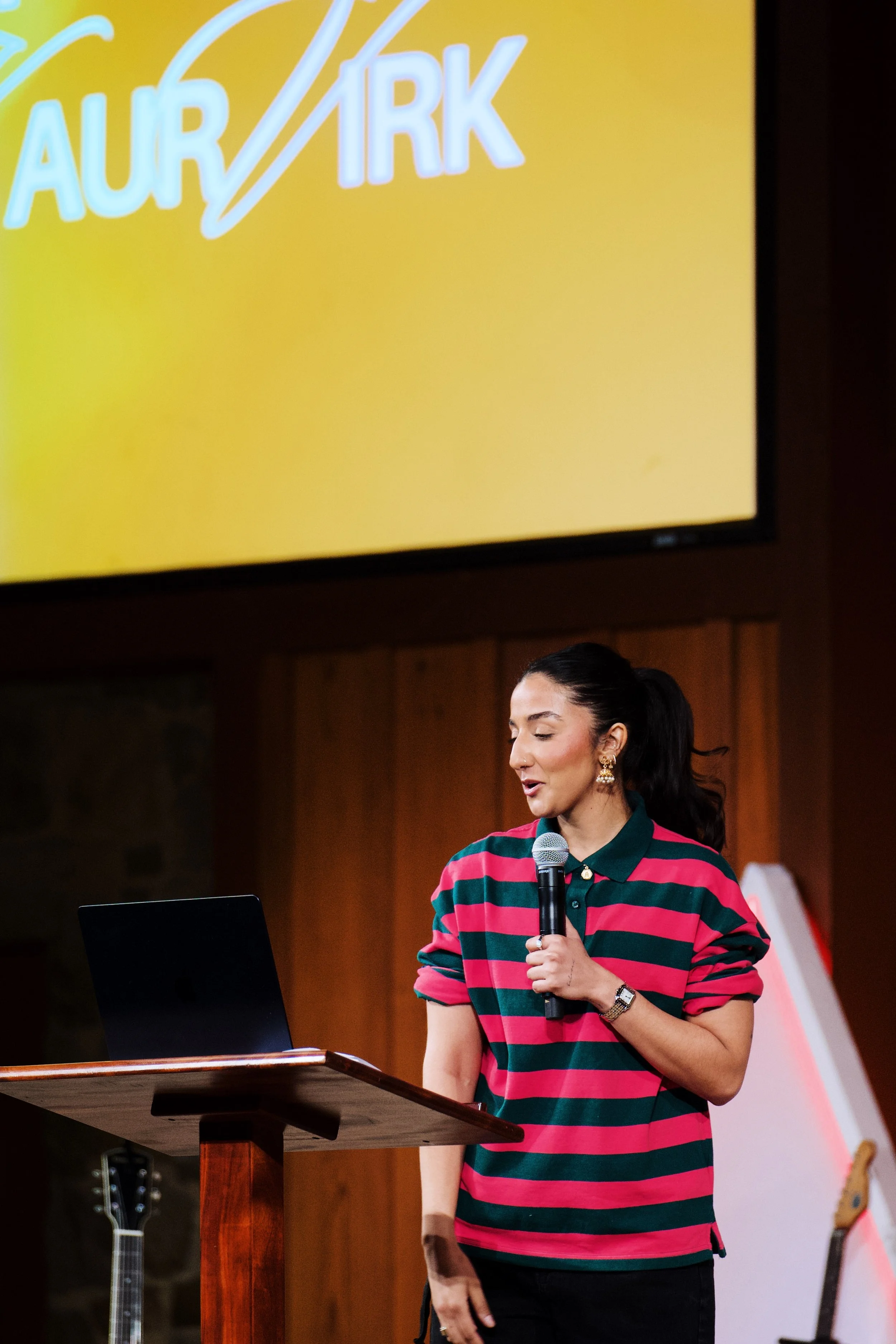 A woman with dark hair tied back, wearing gold earrings and a red and green striped polo shirt, standing at a wooden podium with a laptop in front of her. She is holding a microphone and appears to be speaking or presenting in a room with music instruments, a large screen overhead displaying the words 'AUR' and 'ARK' in bright letters.