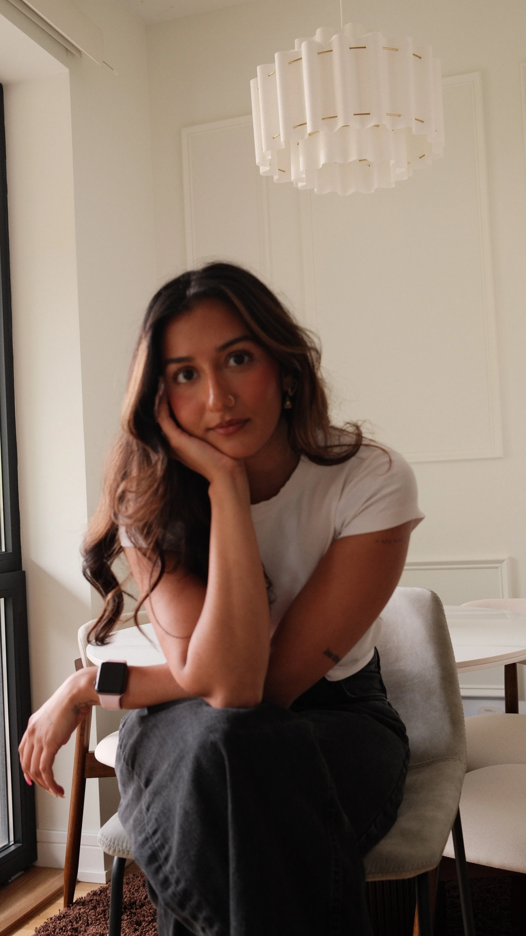 A woman with wavy dark hair, wearing a white T-shirt and black jeans, sitting on a beige chair in a modern, minimally decorated room. She rests her chin on her hand and looks at the camera with a relaxed expression. There is a white ceiling lamp and a white paneled wall in the background.