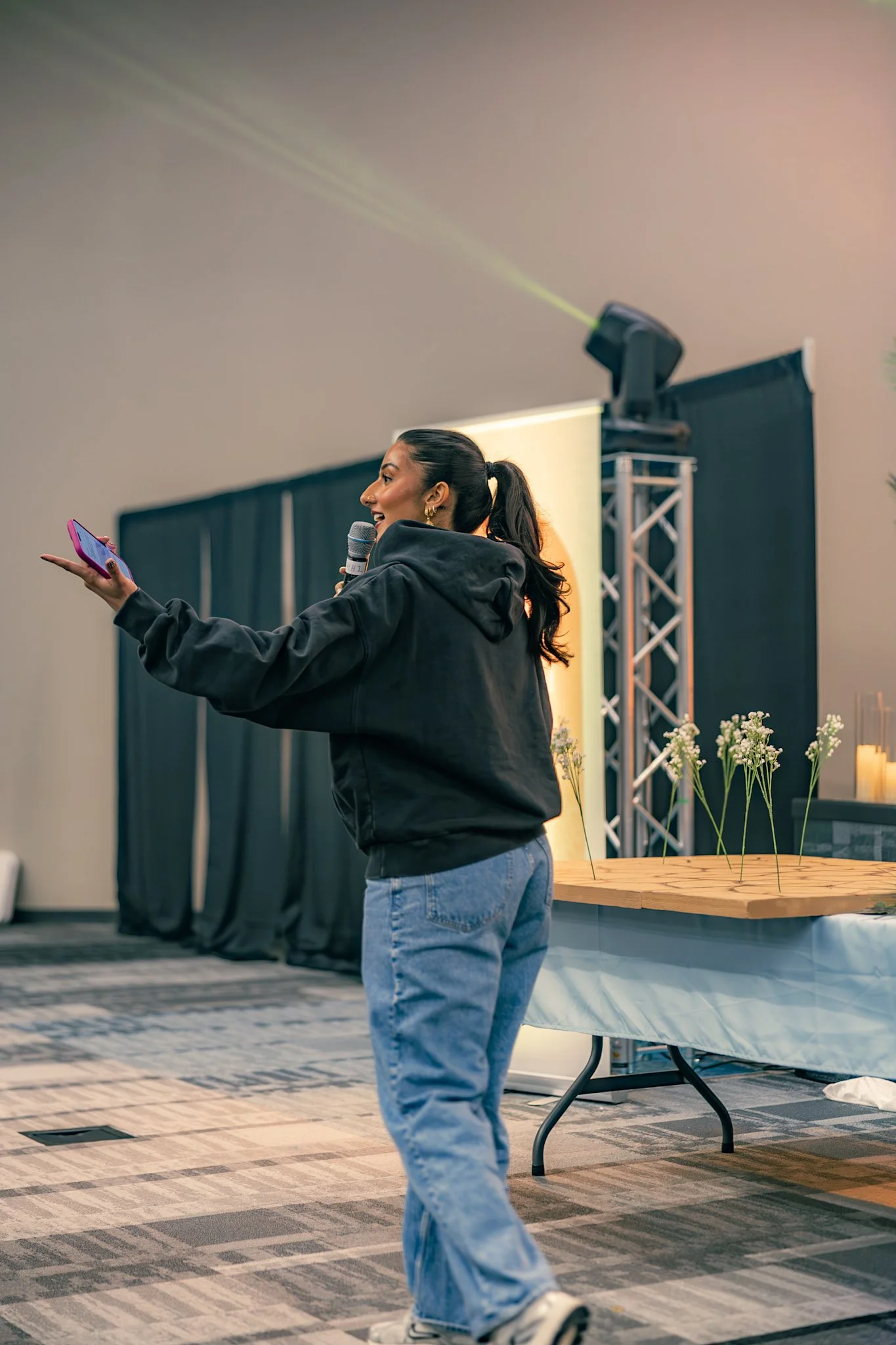 A woman with dark hair tied back, wearing a black hoodie and blue jeans, is standing and speaking into a microphone, taking a selfie with her smartphone during an indoor event with a black curtain, stage lights, and a table with flowers in the background.