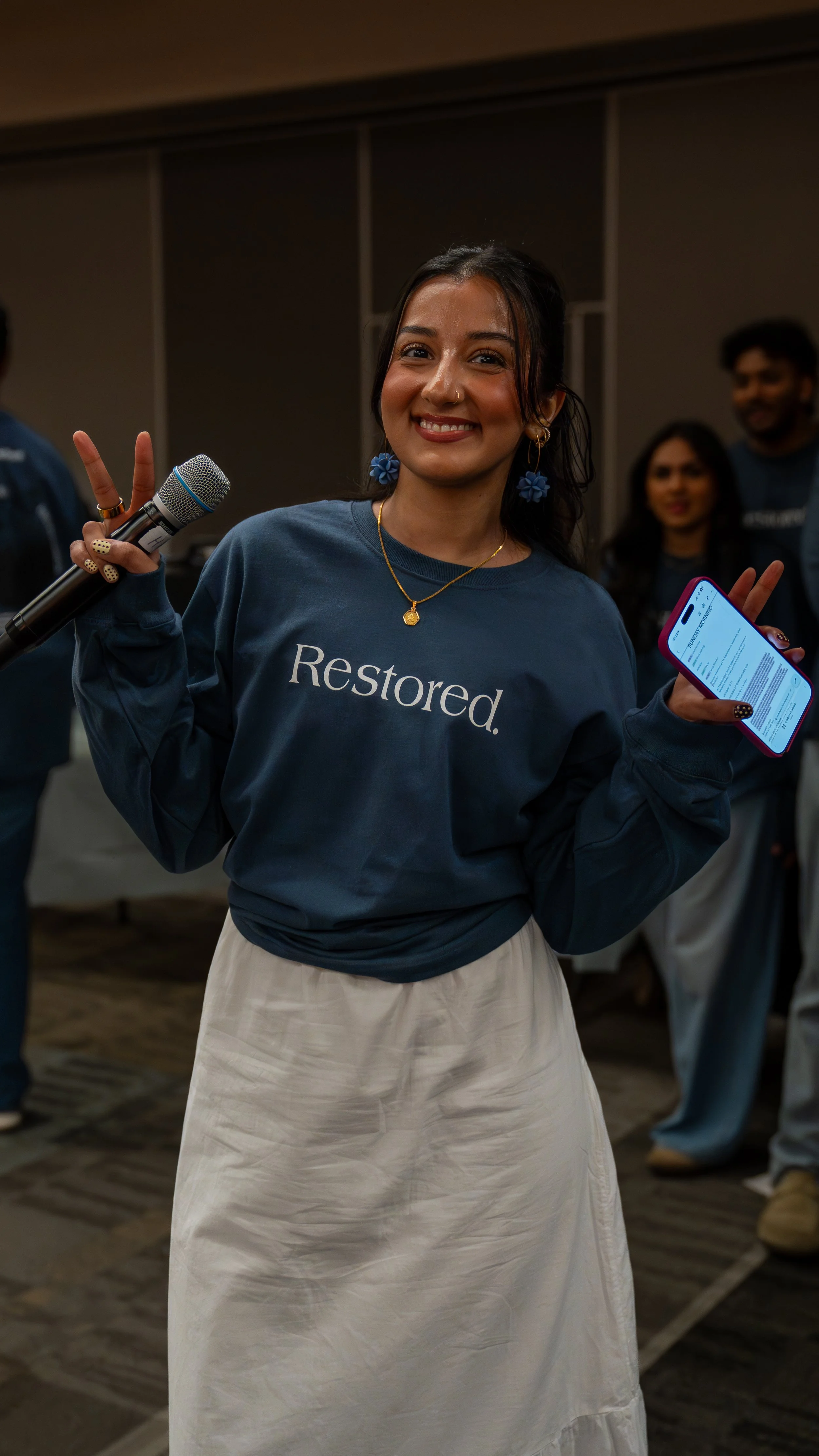 A smiling woman wearing a blue sweatshirt with the word 'Restored.' printed on it, holding a microphone in one hand and a phone in the other. She is making a peace sign with her fingers, standing indoors with a group of people in the background.