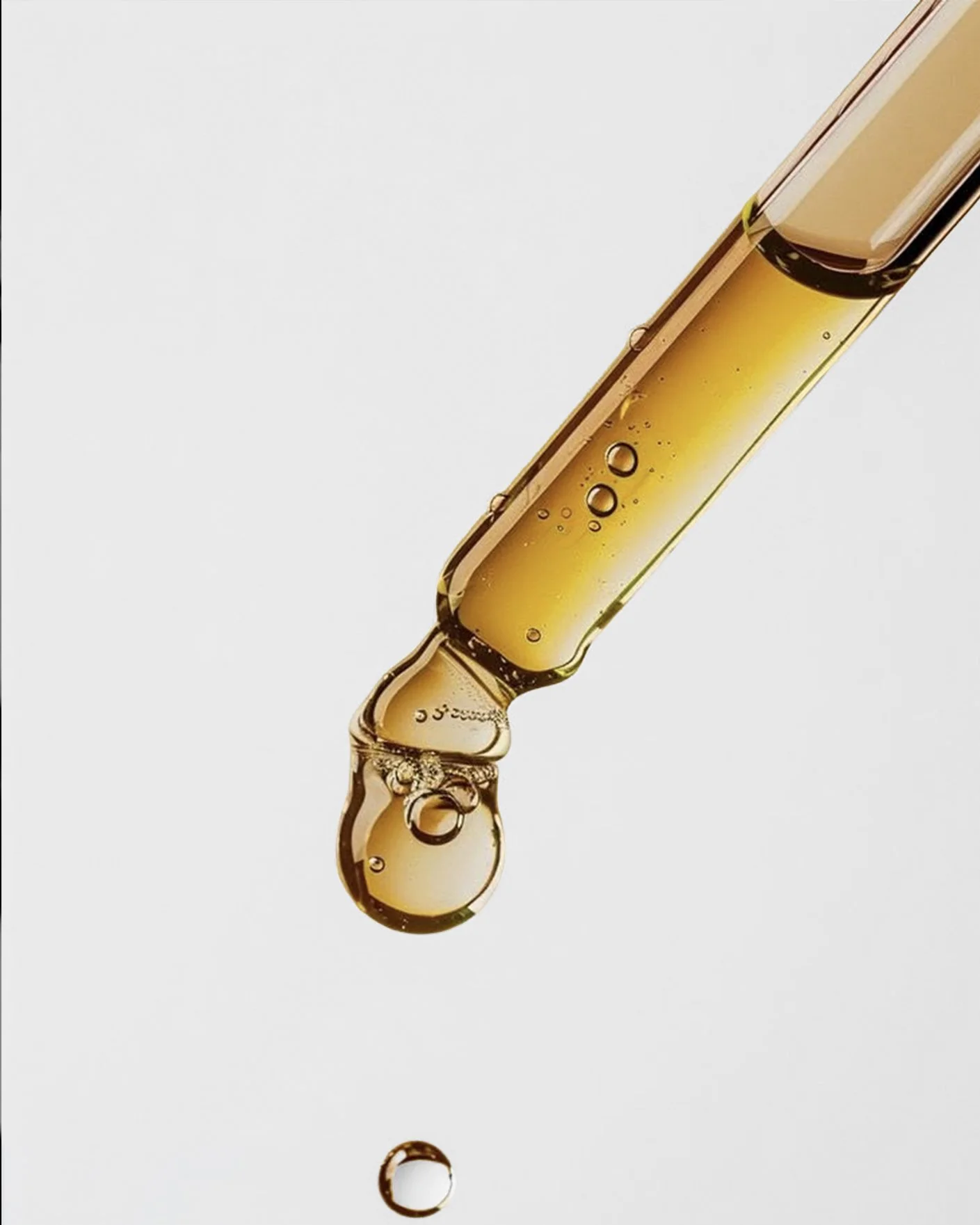 A close-up of a dropper with a golden liquid, possibly oil or serum, with a drop forming at the tip against a plain white background.