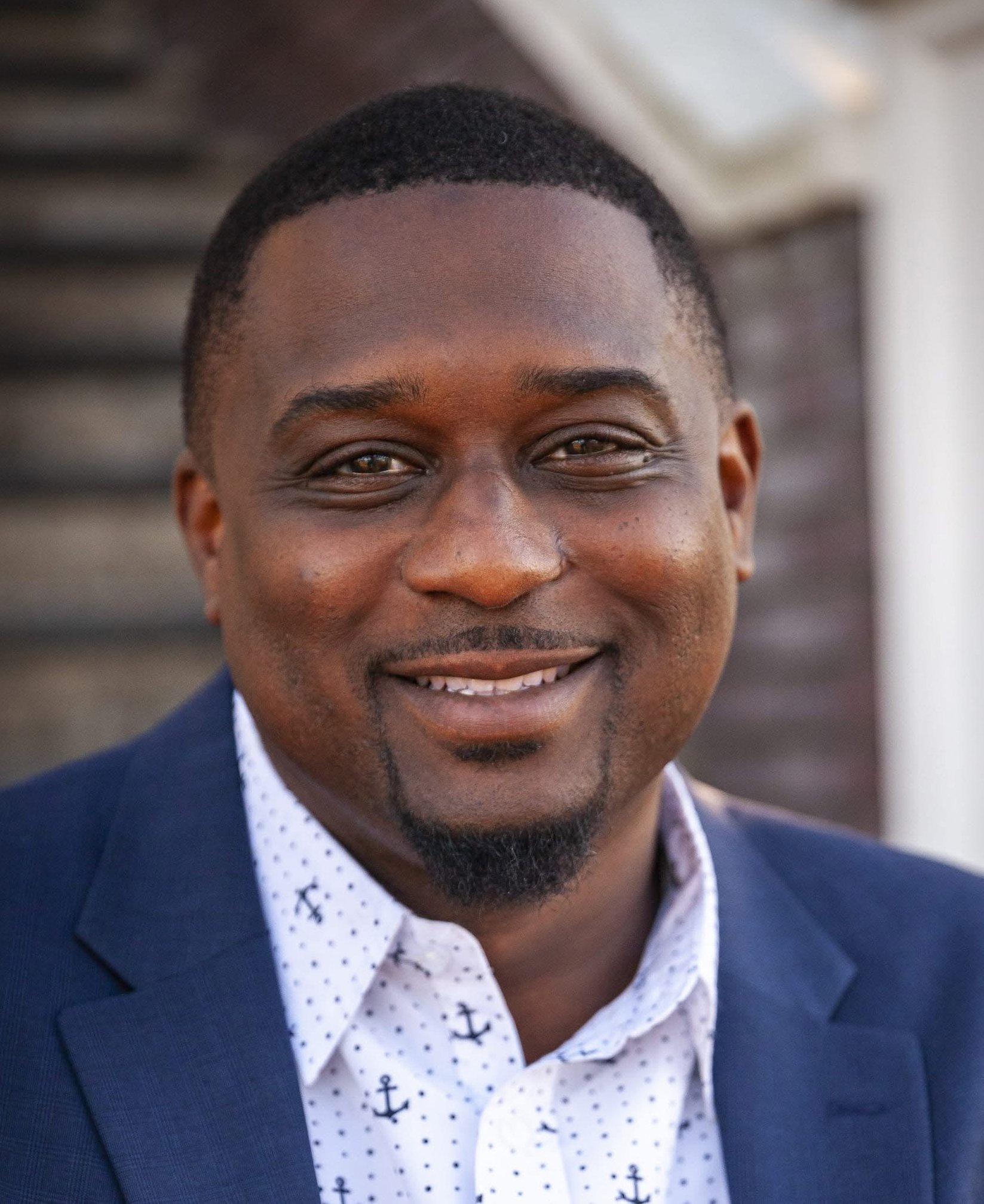 Close-up portrait of a smiling young African American man in a navy blazer and white shirt with black anchors pattern, outdoors with blurred background.
