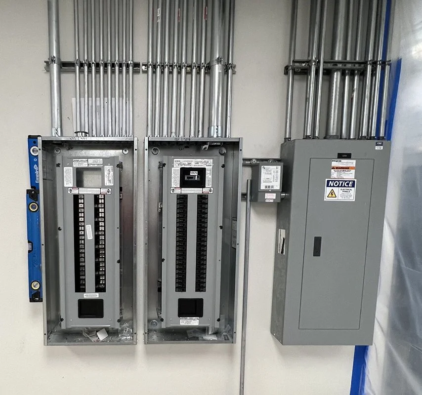 Electrical panel room with two open breaker boxes and a gray electrical cabinet, all mounted on a white wall with metal conduit pipes overhead.